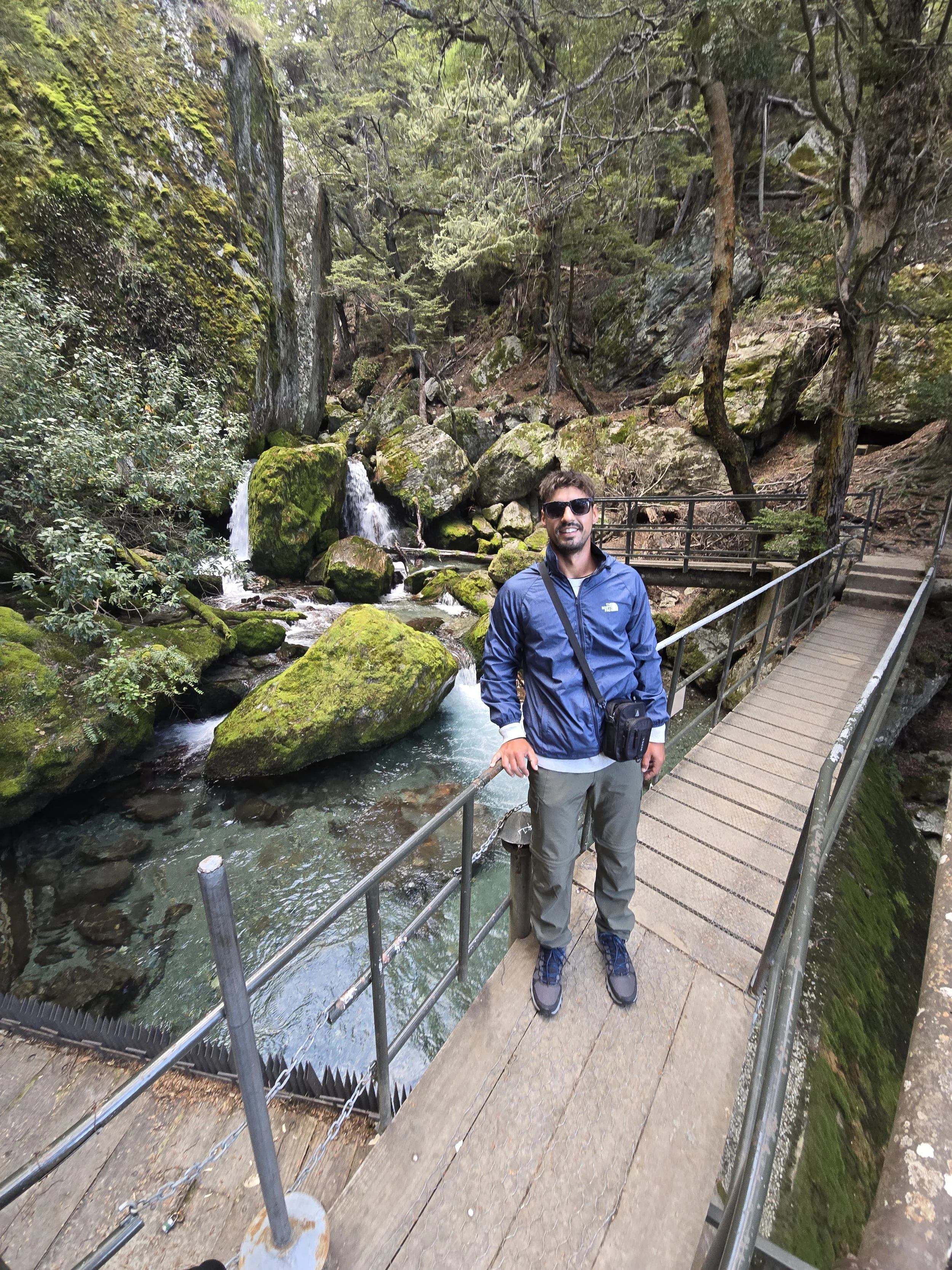 A man wearing sunglasses, a blue jacket, and gray pants is standing on a bridge in a forested area next to a flowing stream with moss-covered rocks. Tall trees and large rocks surround the scene.
