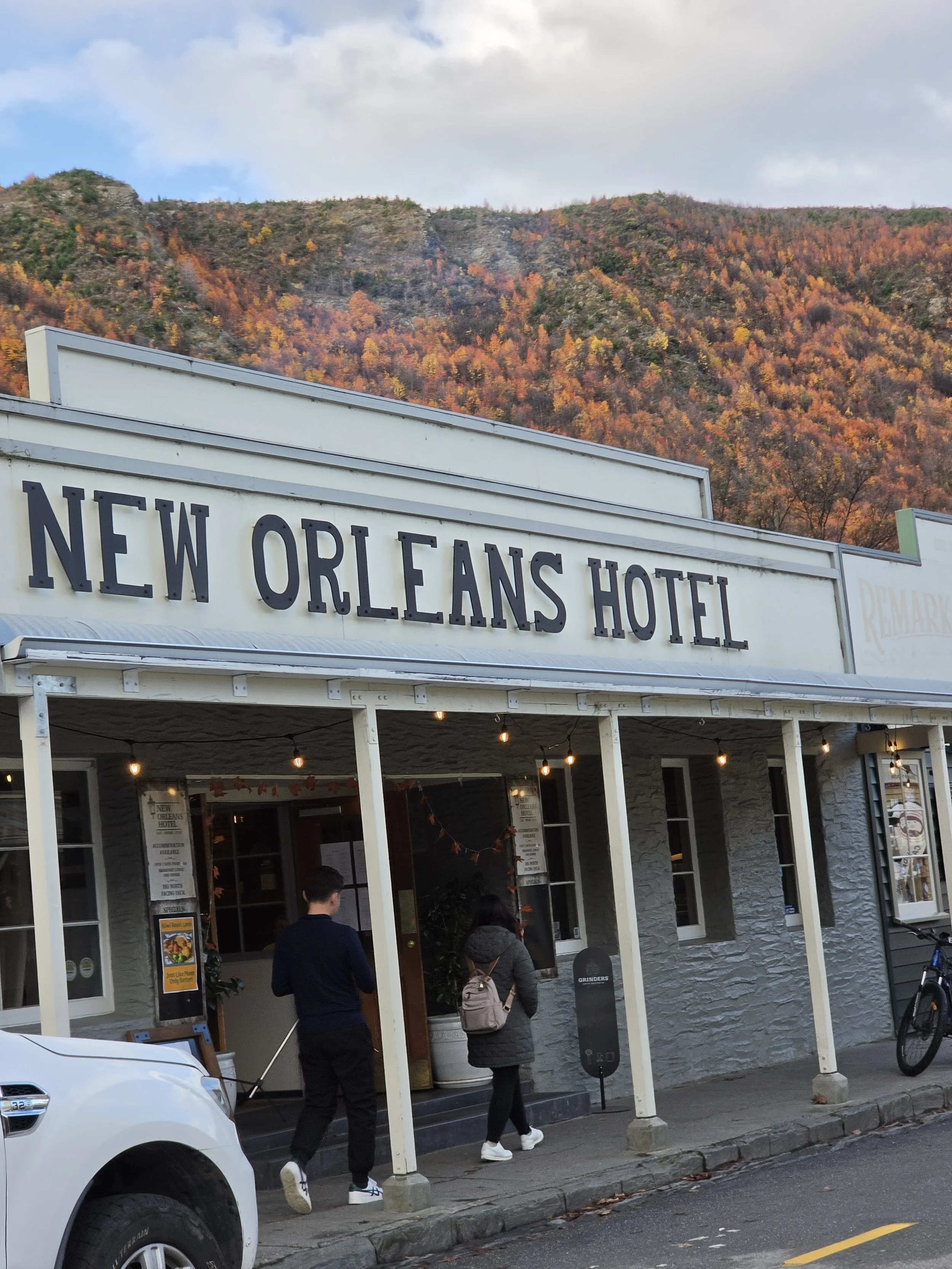 The image shows the exterior of the New Orleans Hotel with a mountain covered in autumn trees in the background. People are walking in and out of the hotel, and some are standing near the entrance. There is a car parked on the street.