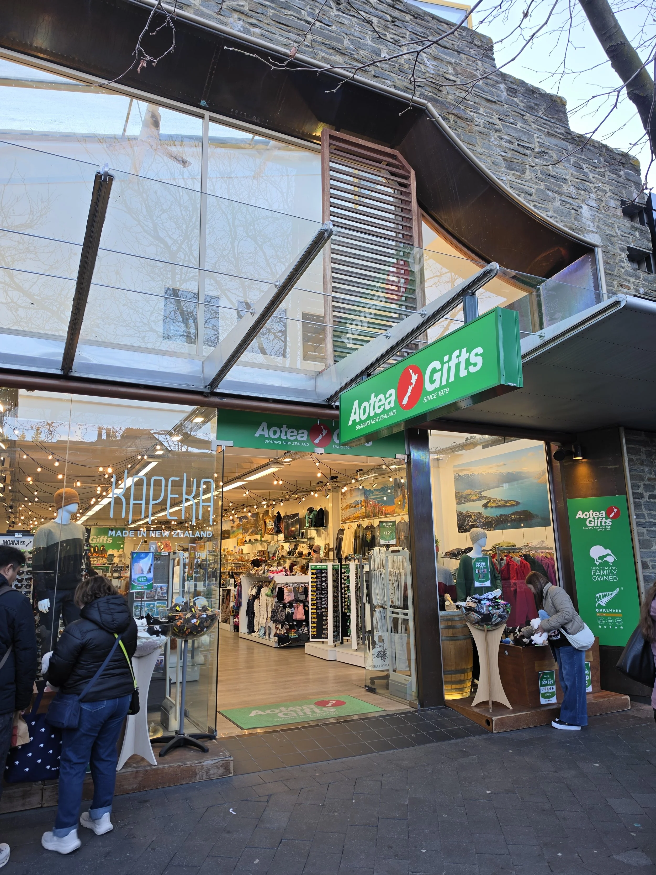 People shopping outside a gift store with a glass storefront, displaying clothing and accessories, with a sign that reads "Aotea Gifts" in green and red.