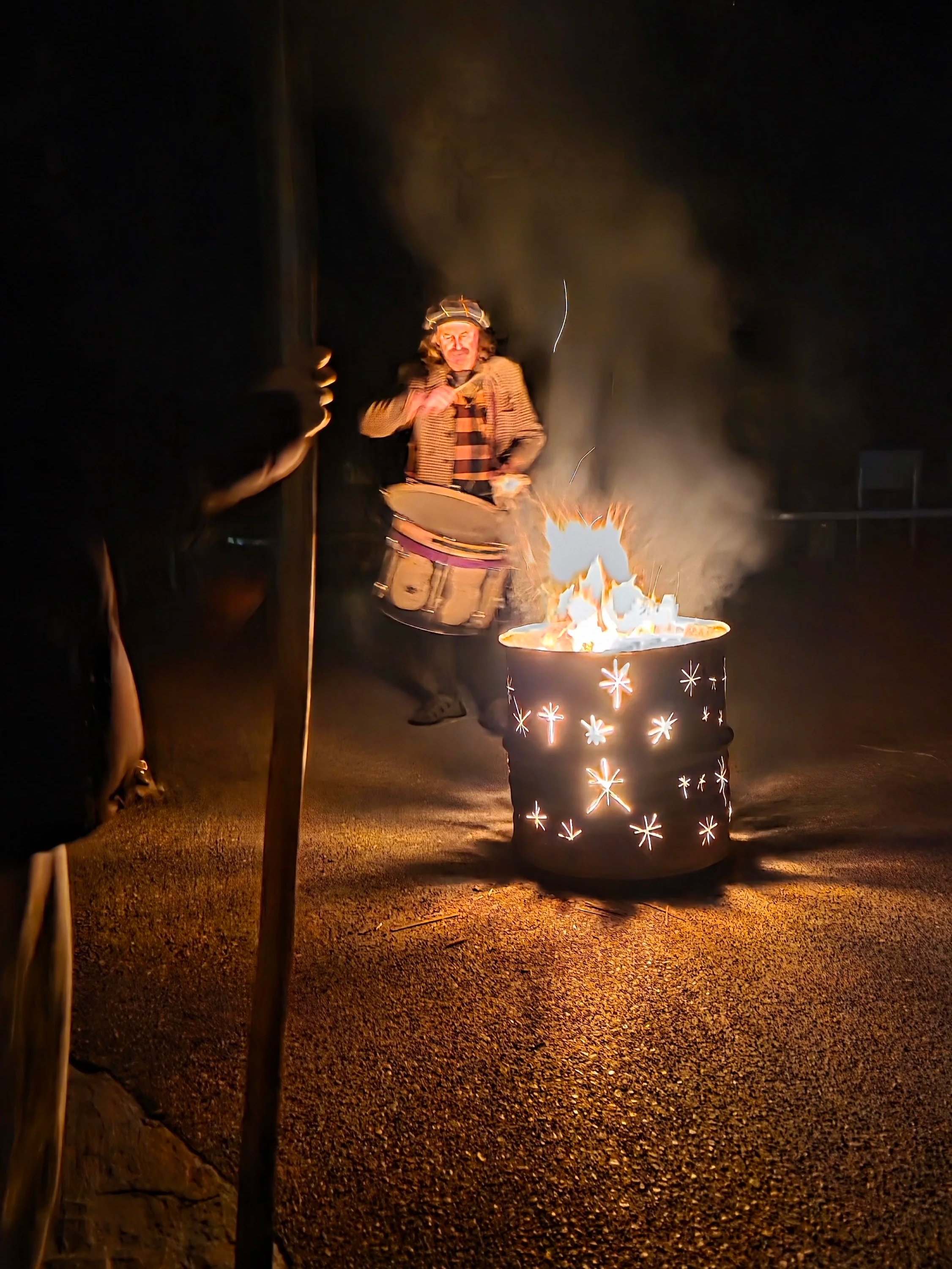 A person standing outside at night near a lit fire in a metal drum with star and snowflake patterns, holding a drum and wearing a hat, with smoke in the background.