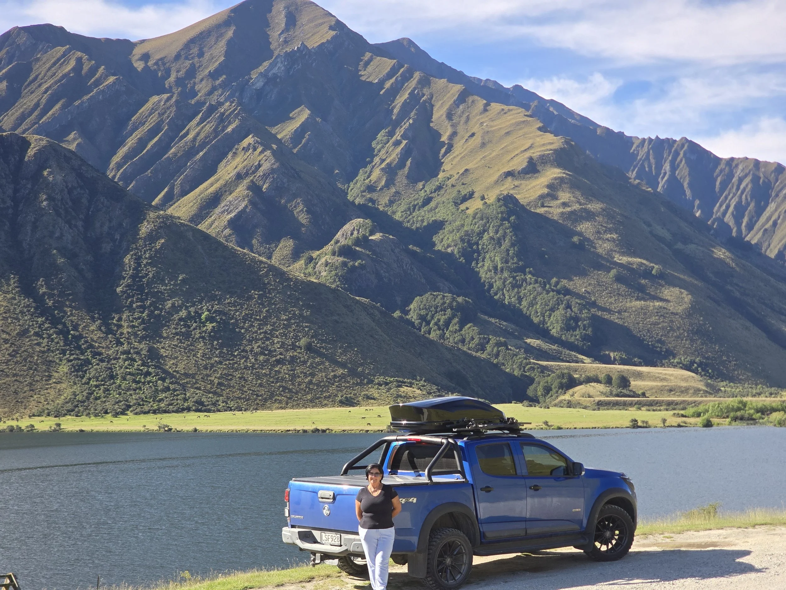 A woman standing next to a blue pickup truck with a mountain lake and green mountains in the background.