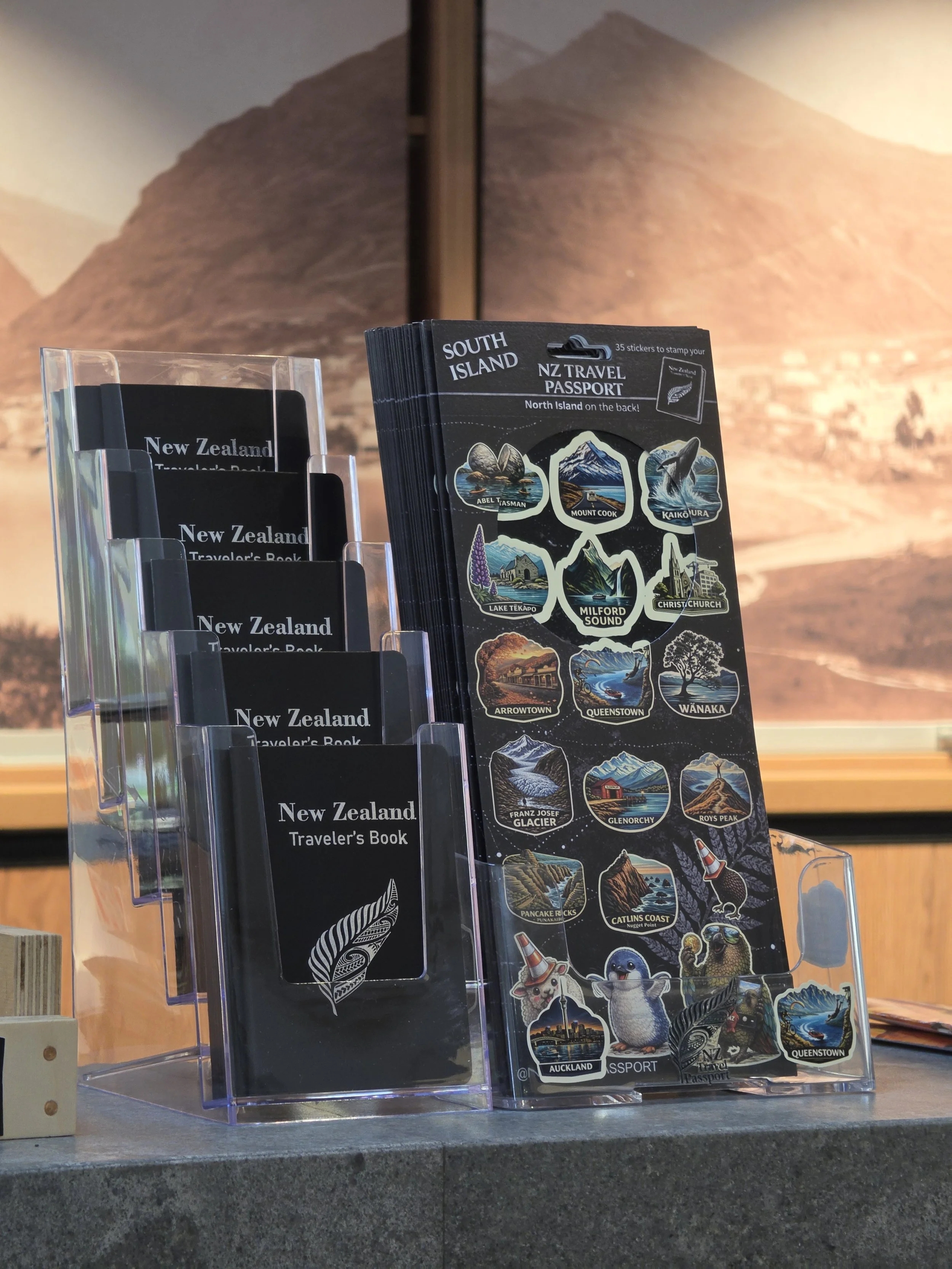 Display of New Zealand travel brochures and stickers with images of mountains, lakes, animals, and landmarks, set against a background of a mountain landscape mural.