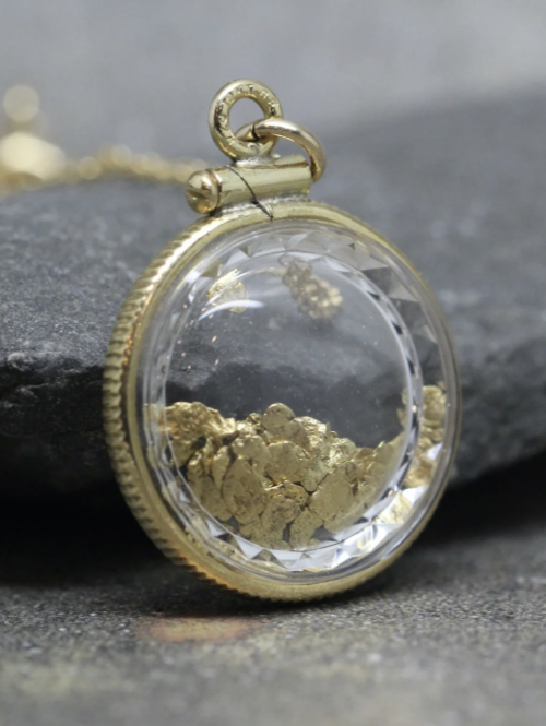 Round glass pendant with gold flakes inside, attached to a gold chain, resting on a dark grey surface.