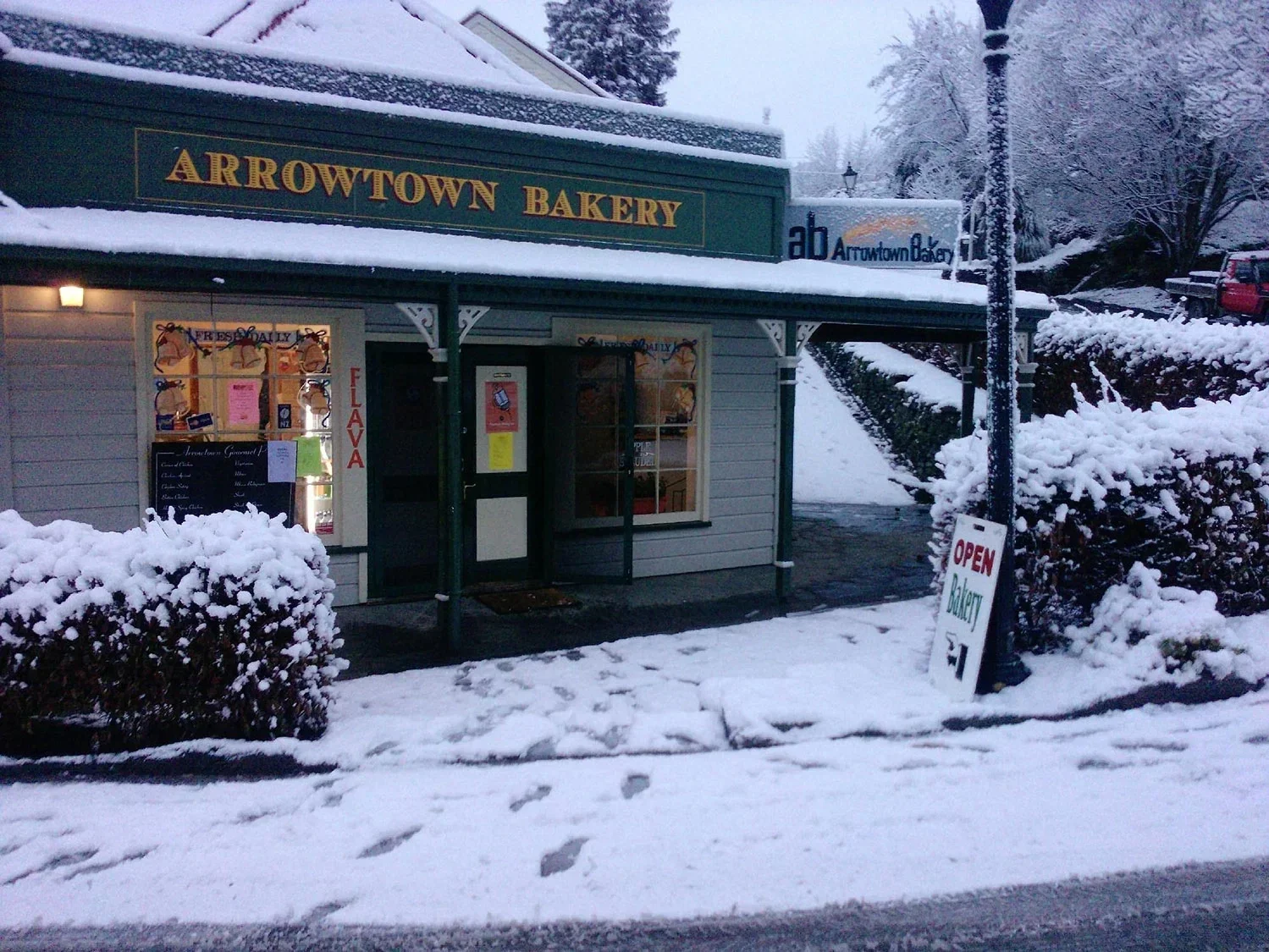 Small bakery named Arrowtown Bakery, with a snow-covered sidewalk and bushes, a signboard and a window display, snowy street with parked cars and trees in the background.