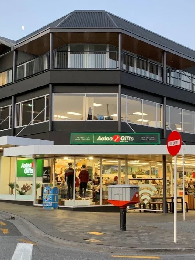 Outside view of a three-story Aotea Gifts store with large glass windows, people inside, a street corner, and signage indicating it is a family-owned business in New Zealand.