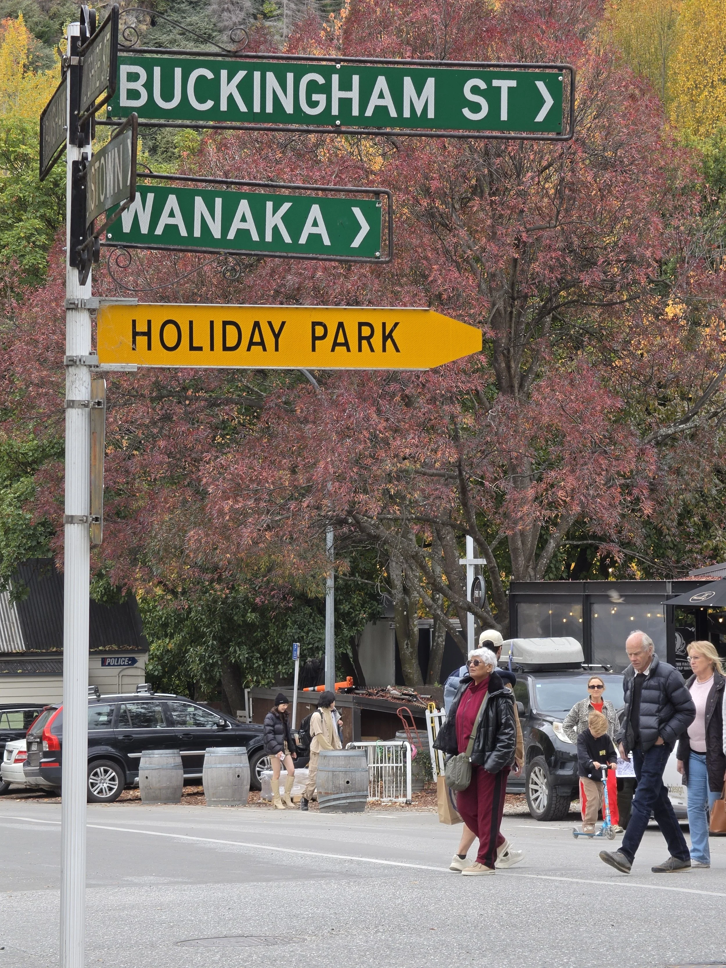 Street scene with people crossing at a crosswalk under street signs for Buckingham Street, Wanaka, and Holiday Park. Vehicles are parked along the street, and trees with autumn foliage are visible in the background.