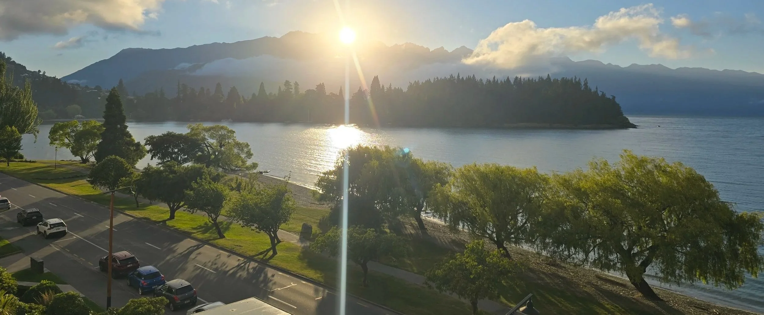 Sun setting over a lake with mountains in the background, trees along the shore, parked cars and a road in the foreground.