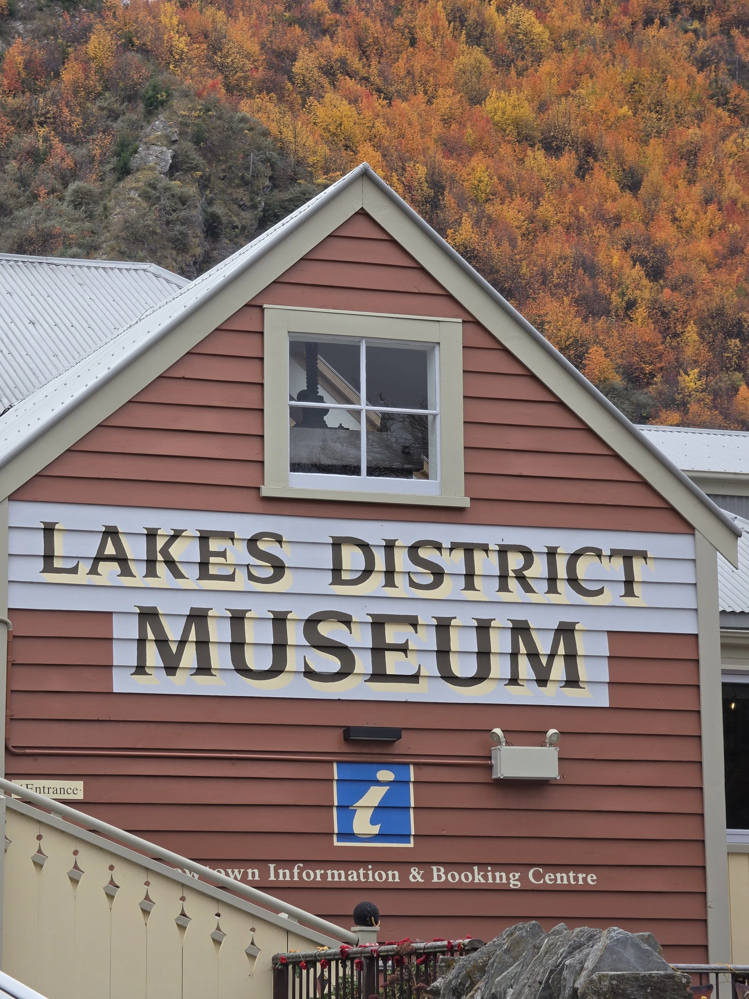 A red building with a sign reading 'Lakes District Museum' and a window on the upper part. Behind the building, there's a mountain with colorful autumn foliage in the background.