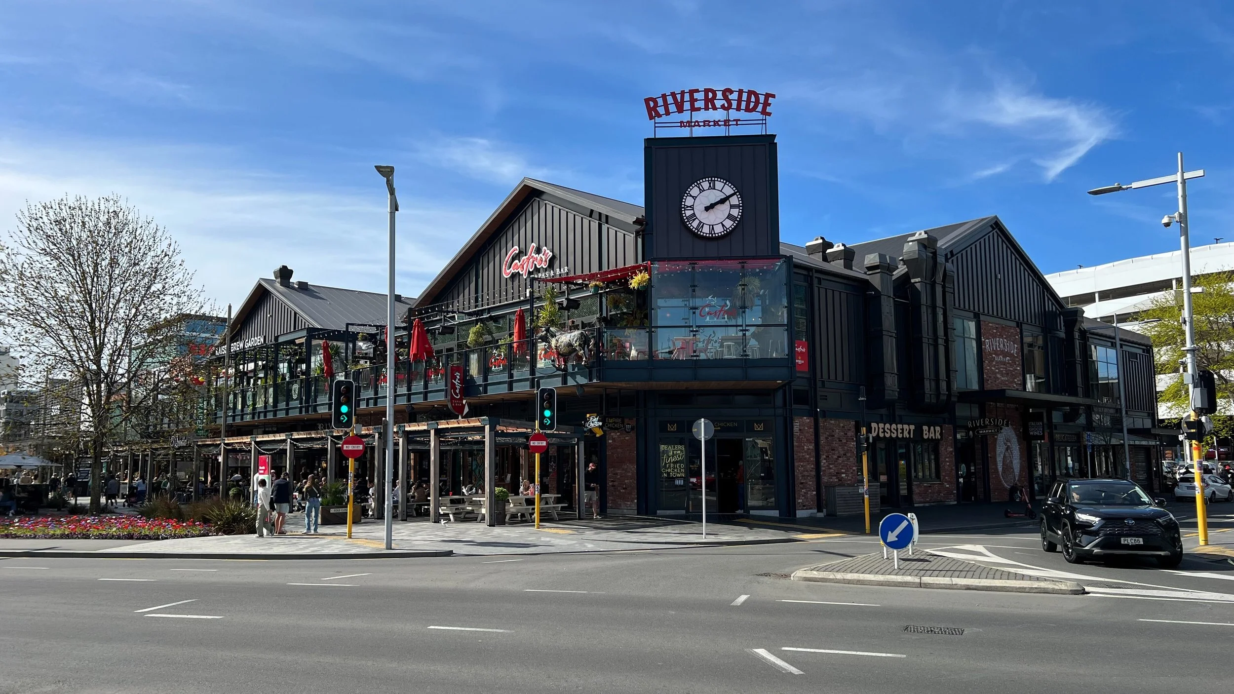 A bustling shopping center with a modern, black building labeled 'Riverside Market' with a clock tower. The building features outdoor seating and signs for a cocktail bar and dessert bar, with people walking and cars at the intersection on a sunny day.
