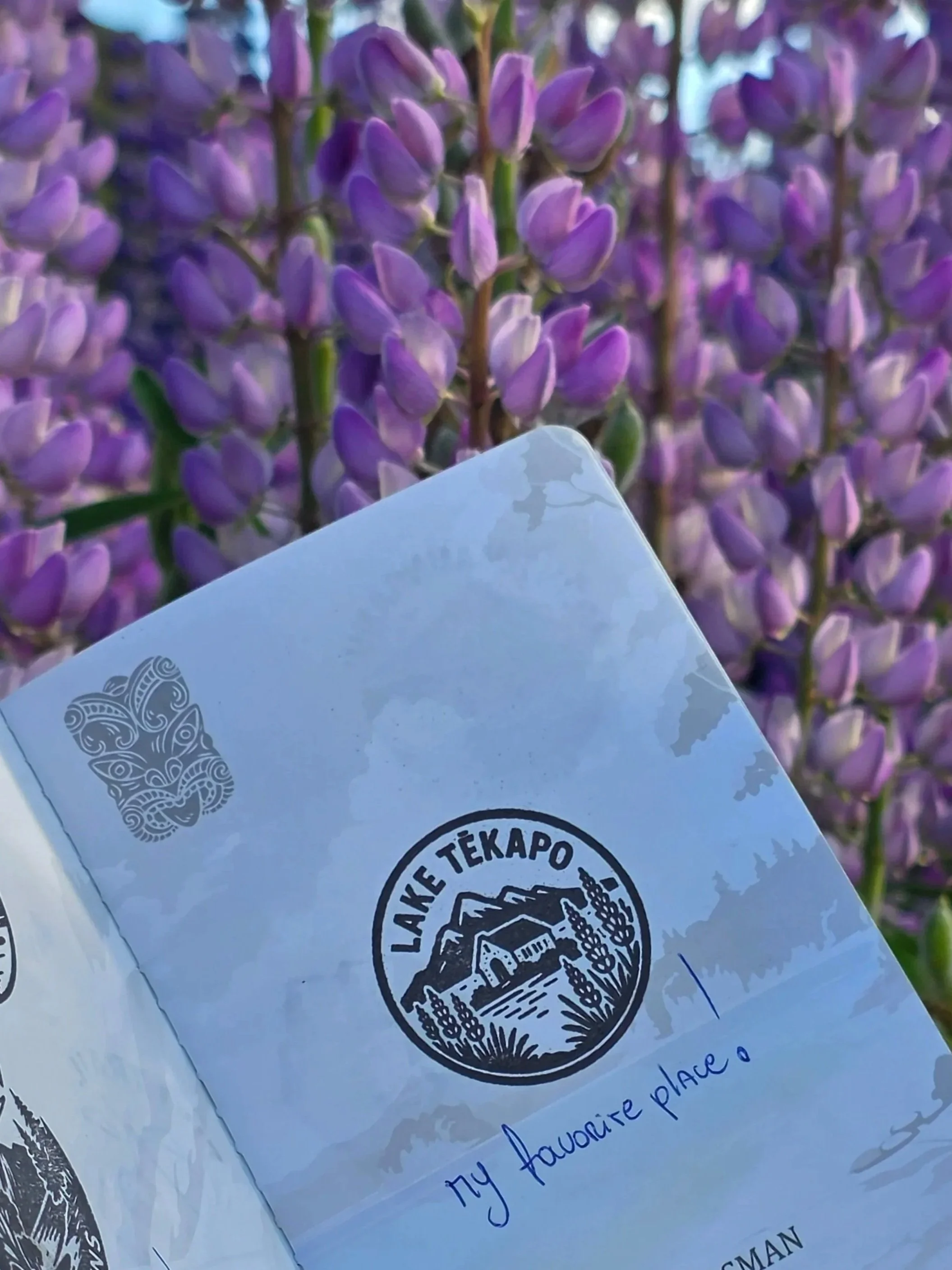 Close-up photo of purple lilac flowers with a white piece of paper in the foreground containing a logo and handwritten text.
