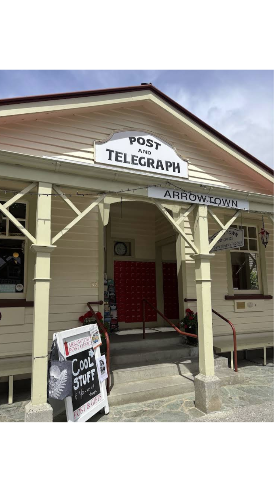 Vintage post office building with signs reading 'Post and Telegraph' and 'Arrowtown Post Office,' featuring a porch with red mailboxes, outdoor signage, and potted flowers.