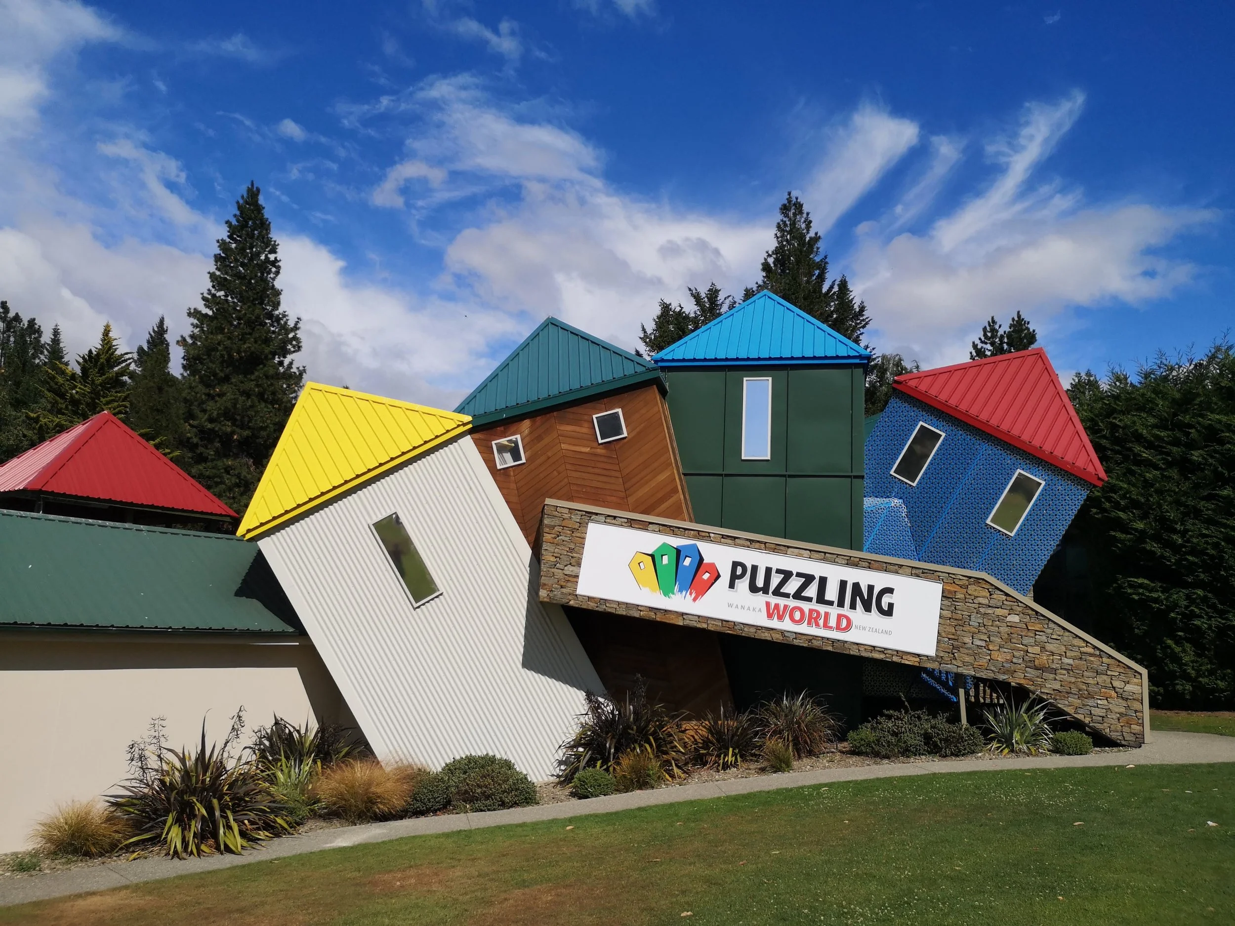Colorful tilted houses with red, yellow, green, and blue roofs behind a sign that reads 'PUZZLING WORLD.' The background features a blue sky with white clouds and tall green trees.