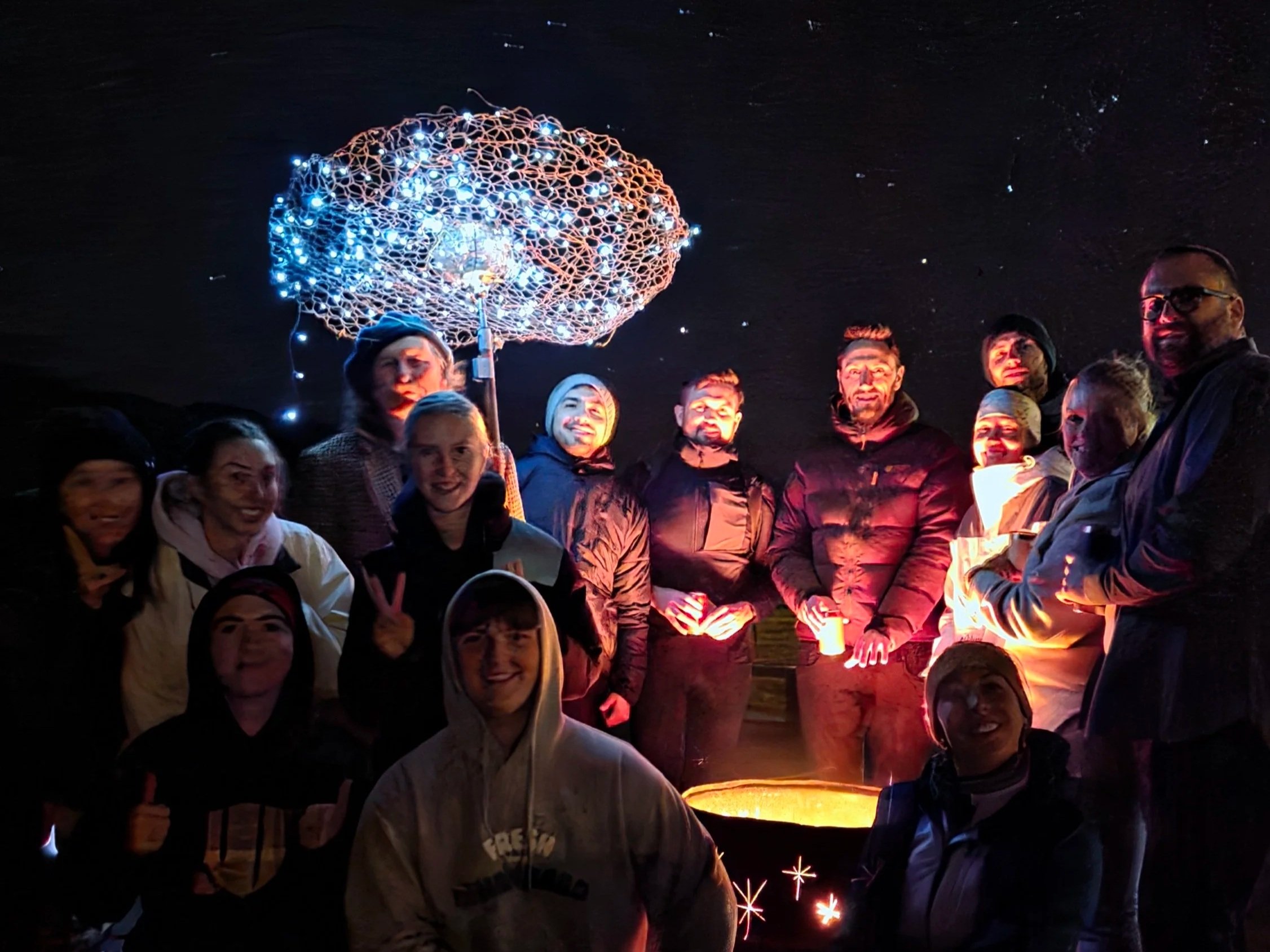 A group of people gathered together at night outdoors, holding sparklers, with a large umbrella-shaped string light setup and a starry night sky in the background.
