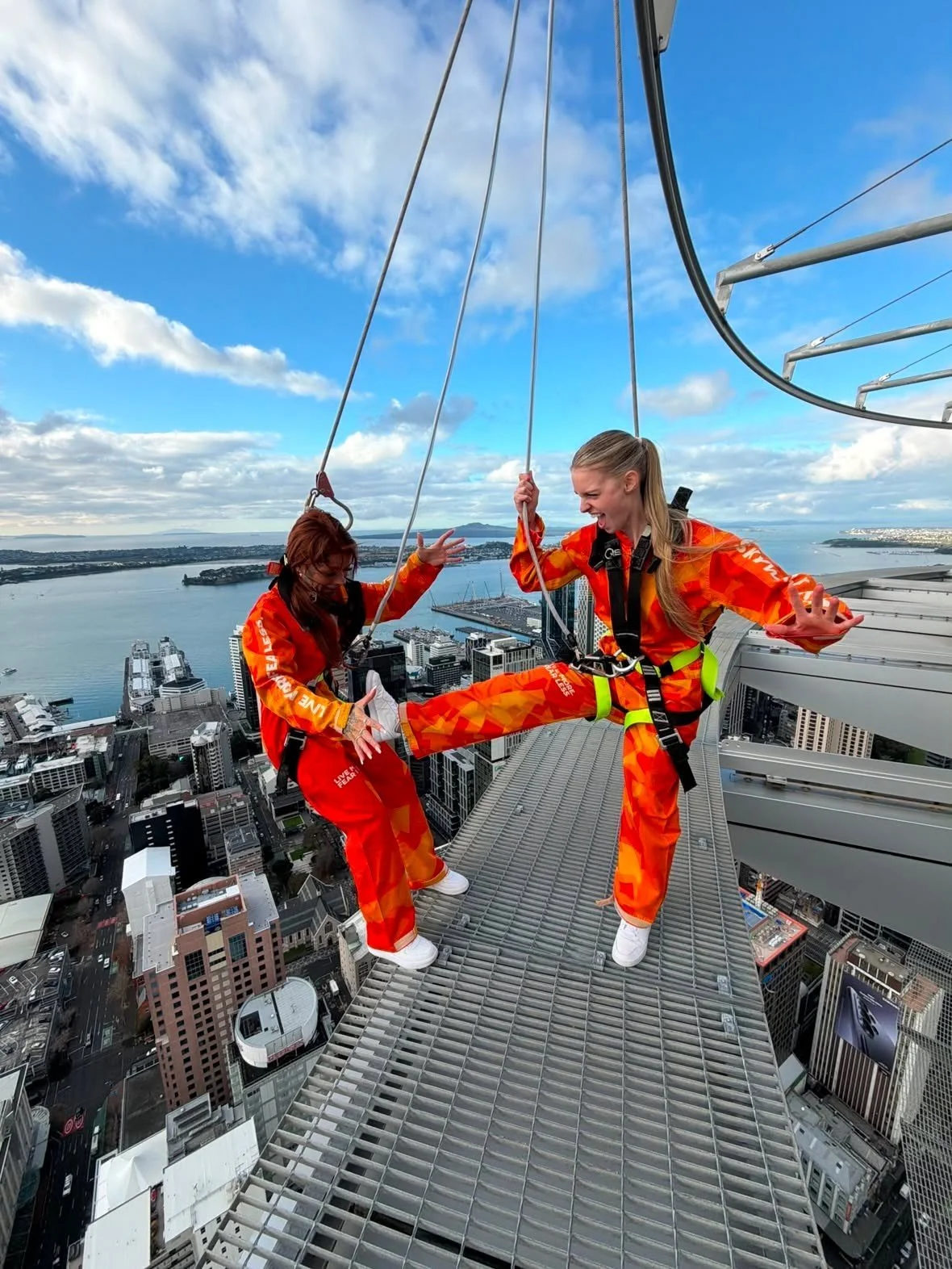 Two women wearing bright orange safety suits are on the ledge of a tall skyscraper, engaging in a bungee jump or similar activity with a city skyline and water in the background.
