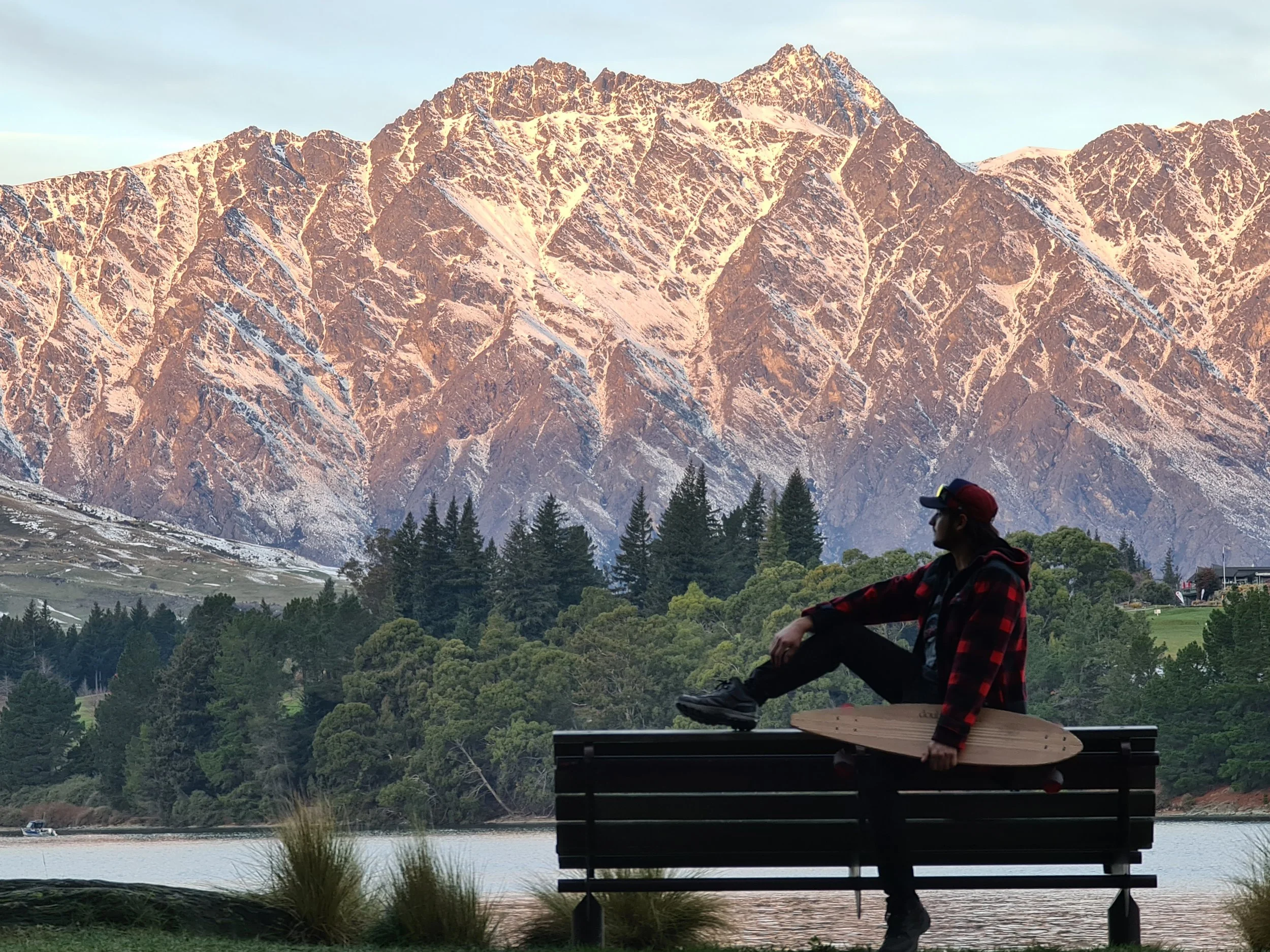A person sitting on a wooden bench near a body of water, holding a skateboard, with a large forested mountain in the background.