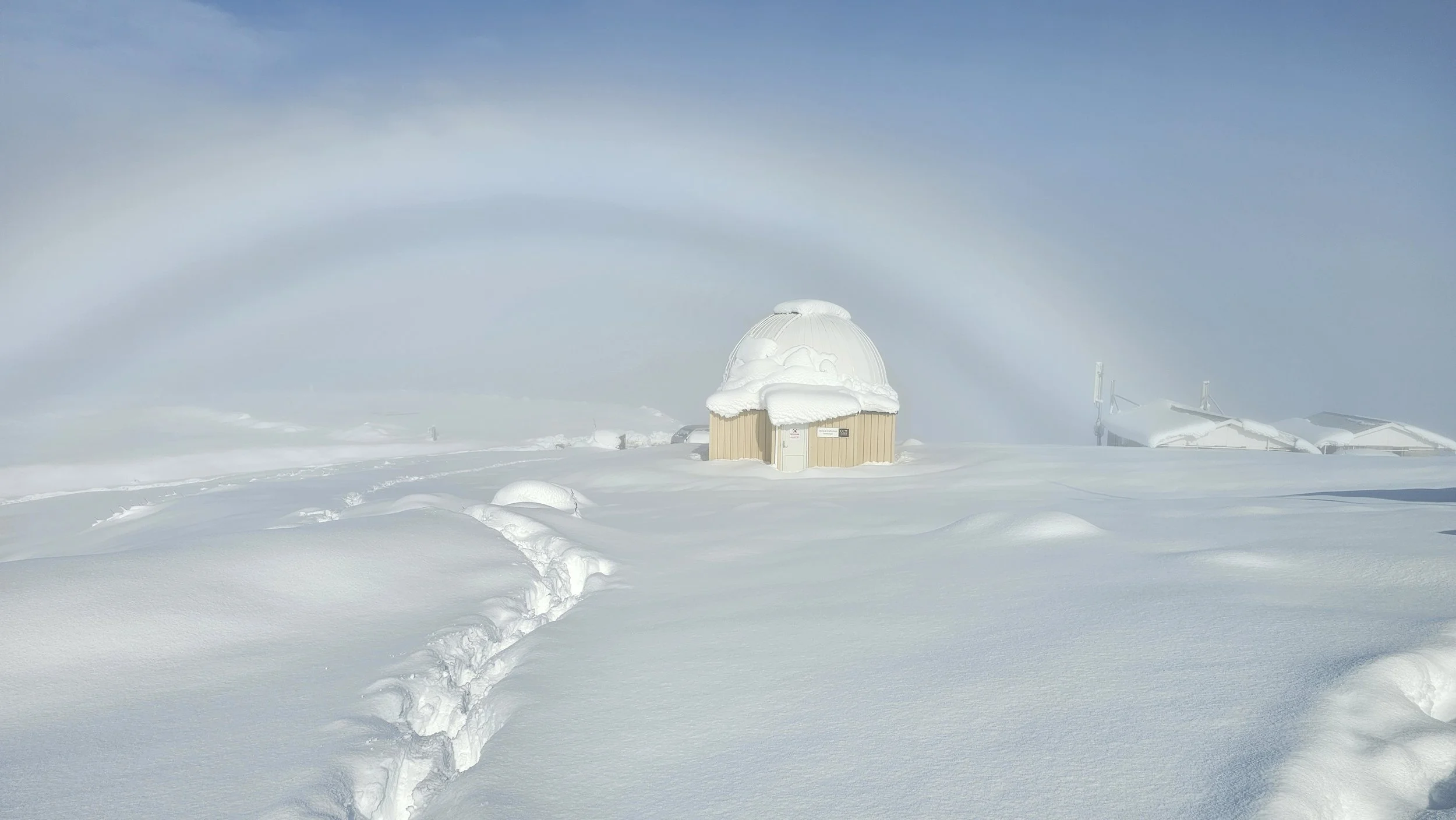 A small weather observatory or shelter covered in snow in a snowy landscape, with tracks leading to it and snow-covered structures in the background under a cloudy sky.