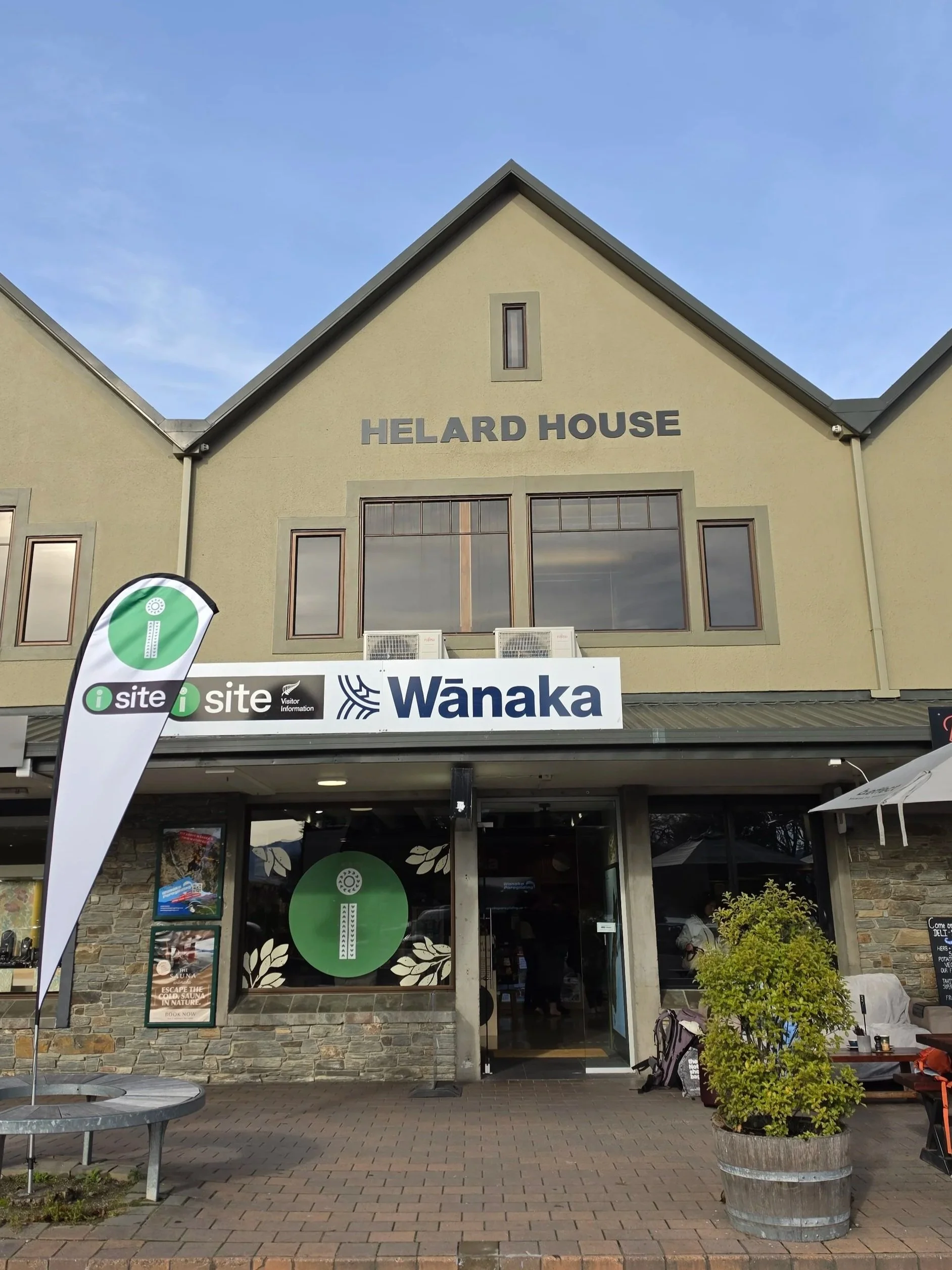 Captivating view of Helard House building with modern design, featuring large windows and beige exterior, adjacent to Wānaka Visitor Information signage, with cloudy sky above and outdoor seating area with a potted plant.