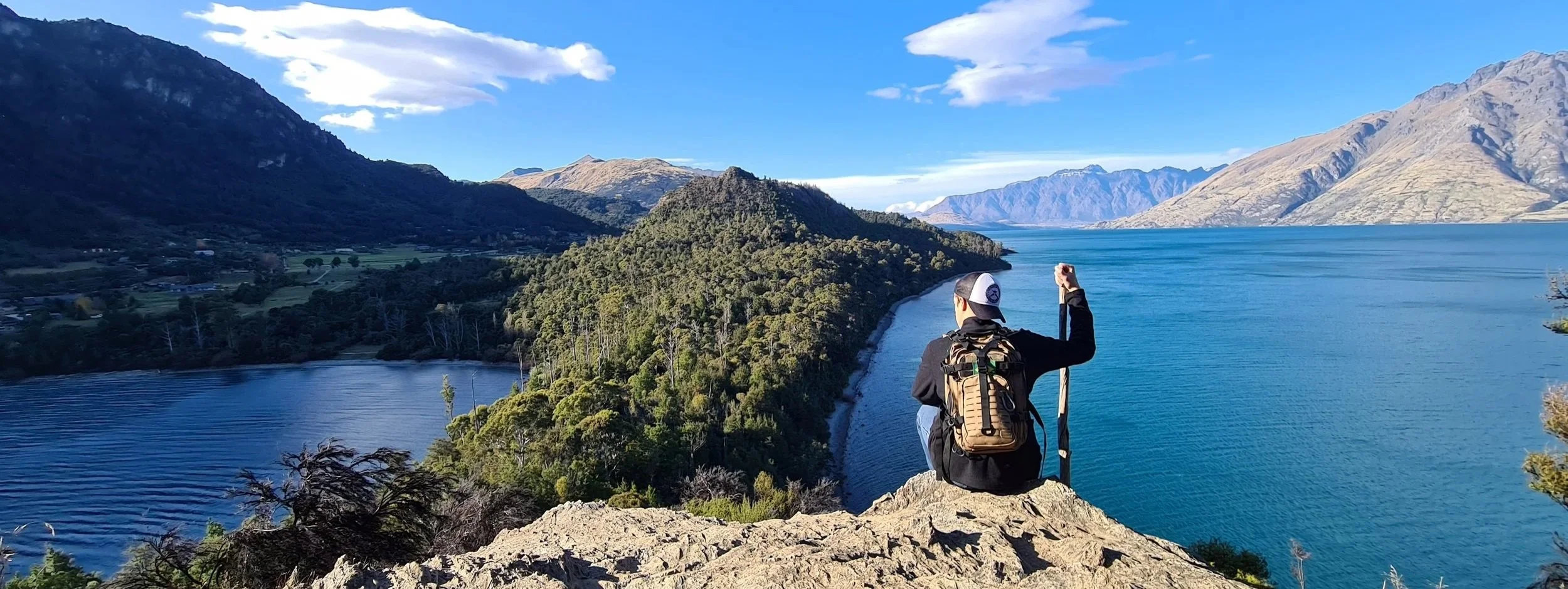 Person sitting on a rock at a scenic lakeside view, holding a walking stick, surrounded by mountains, trees, and a blue sky with clouds.