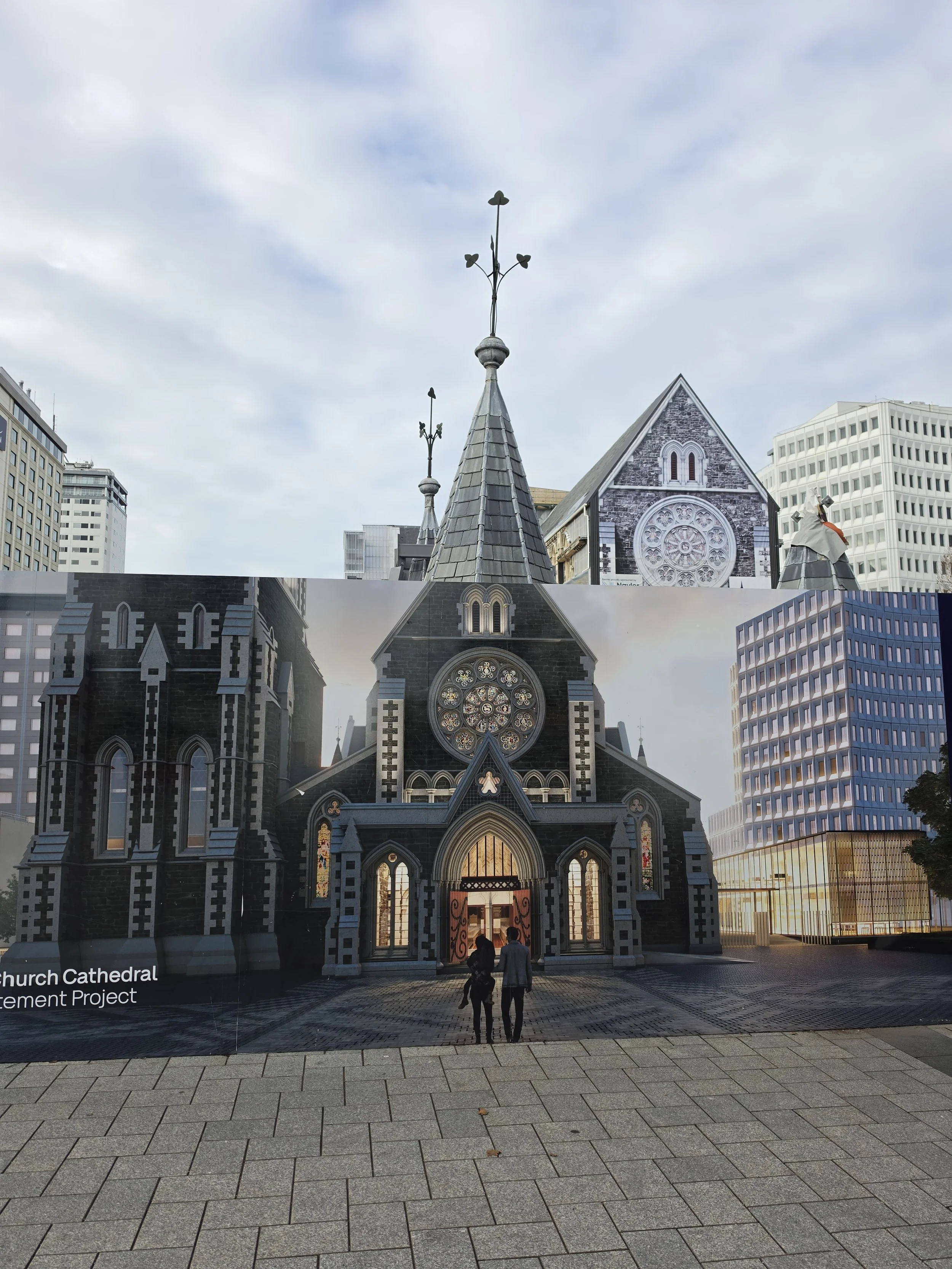A Gothic-style church with a stone facade, large stained glass rose window, three steeples topped with weather vanes, and modern skyscrapers in the background, with a few people walking toward the church entrance.