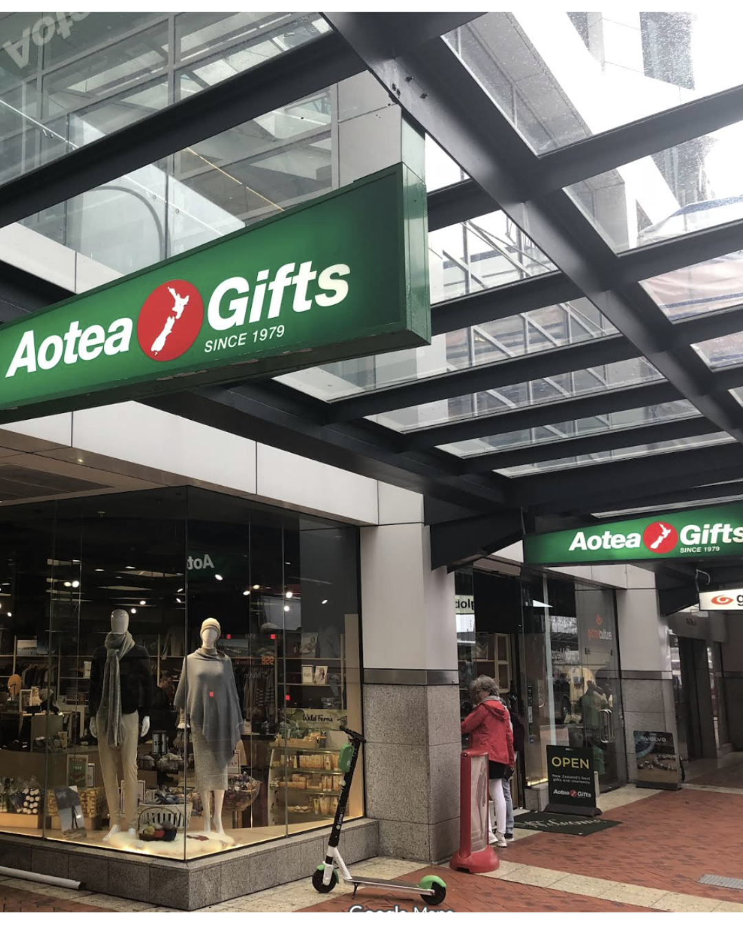 Storefront of Aotea Gifts with mannequins displaying clothing inside and a person near the entrance, located in a shopping area with a scooter parked outside.