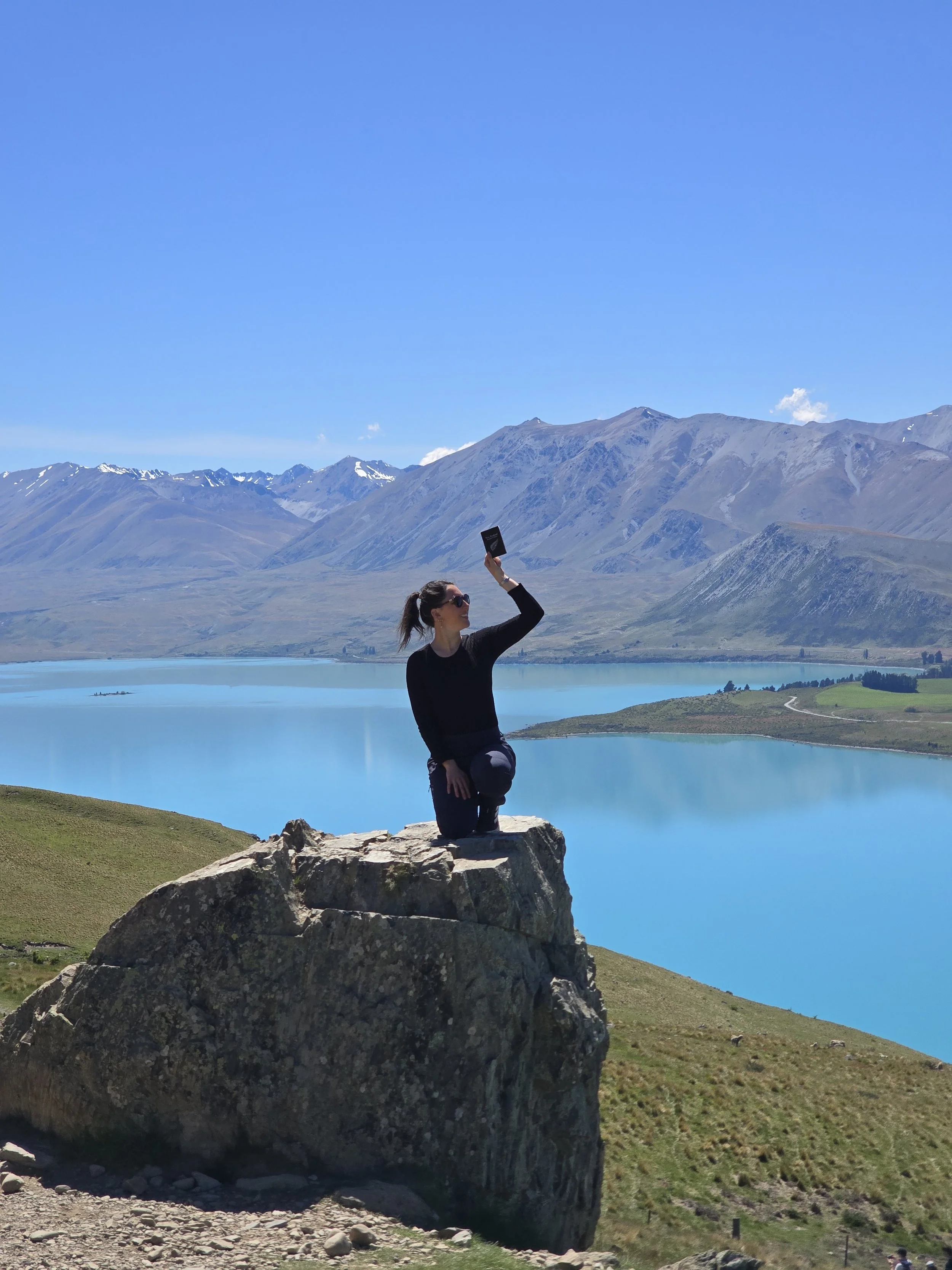 A woman taking a selfie on a rock overlooking a scenic lake with mountains in the background.