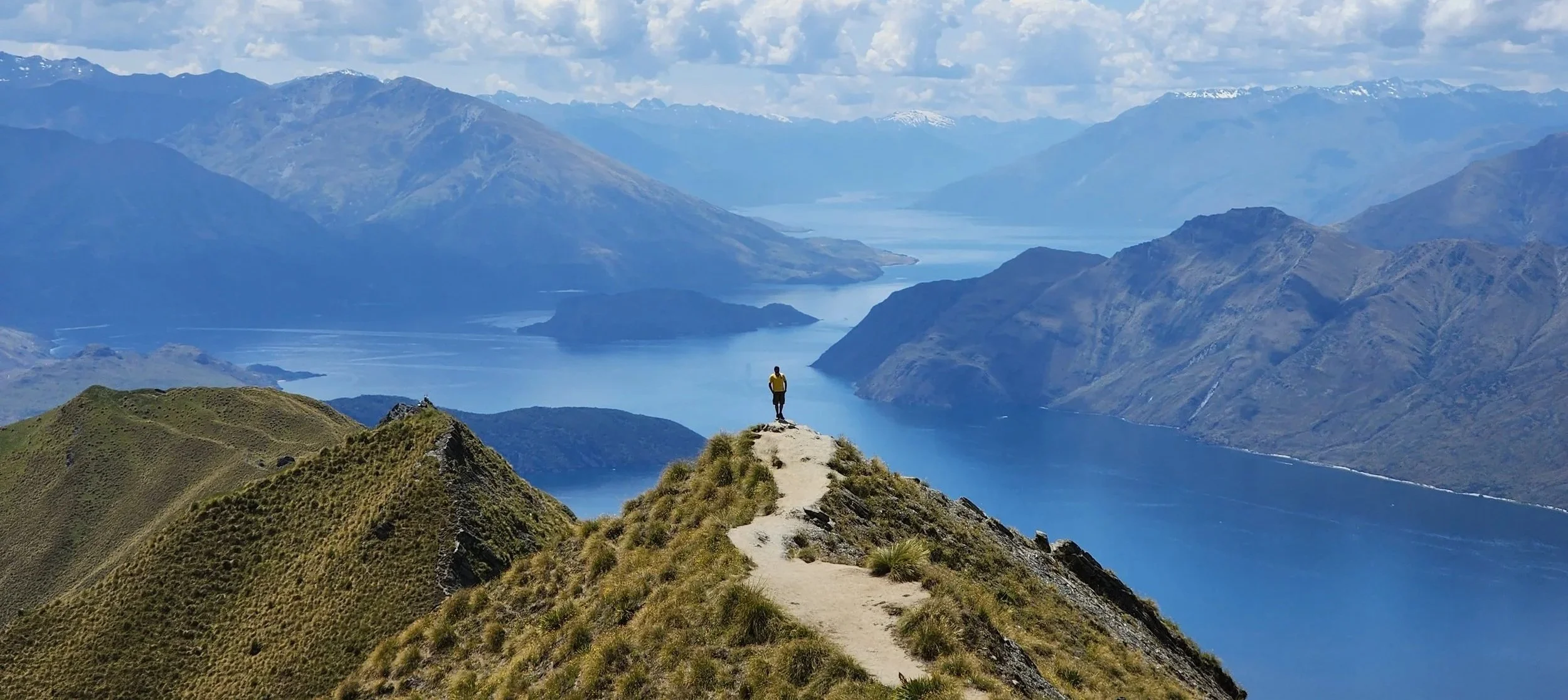A person standing on a mountain ridge overlooking a blue lake and mountain range under a partly cloudy sky.