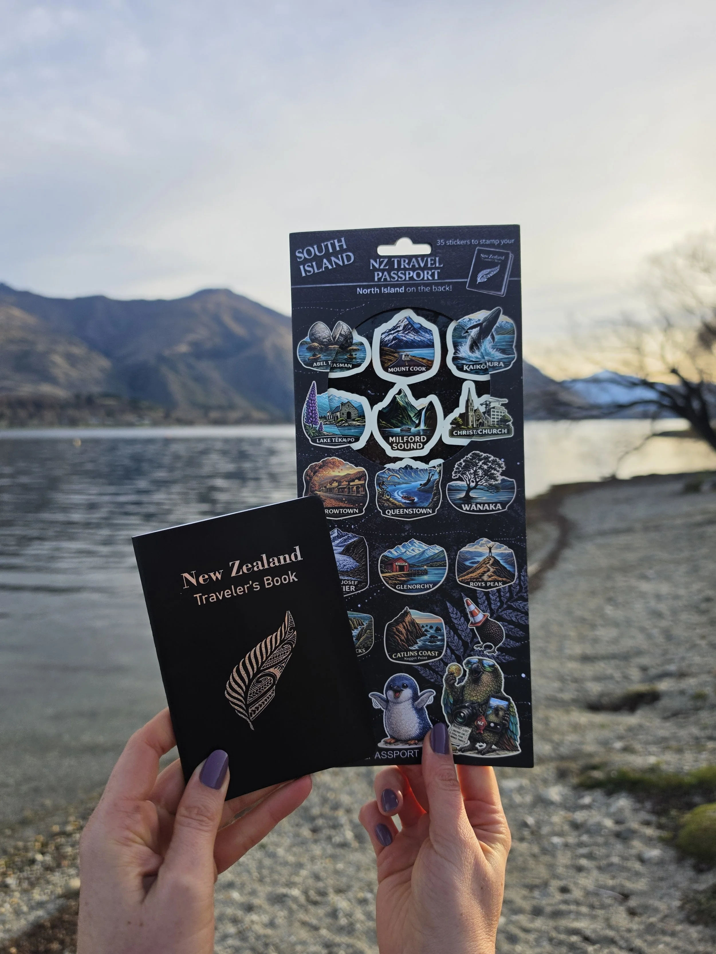 Two hands holding New Zealand travel books and a sticker sheet featuring iconic locations and landmarks of South Island, New Zealand, with mountains and a lake in the background during sunset or sunrise.