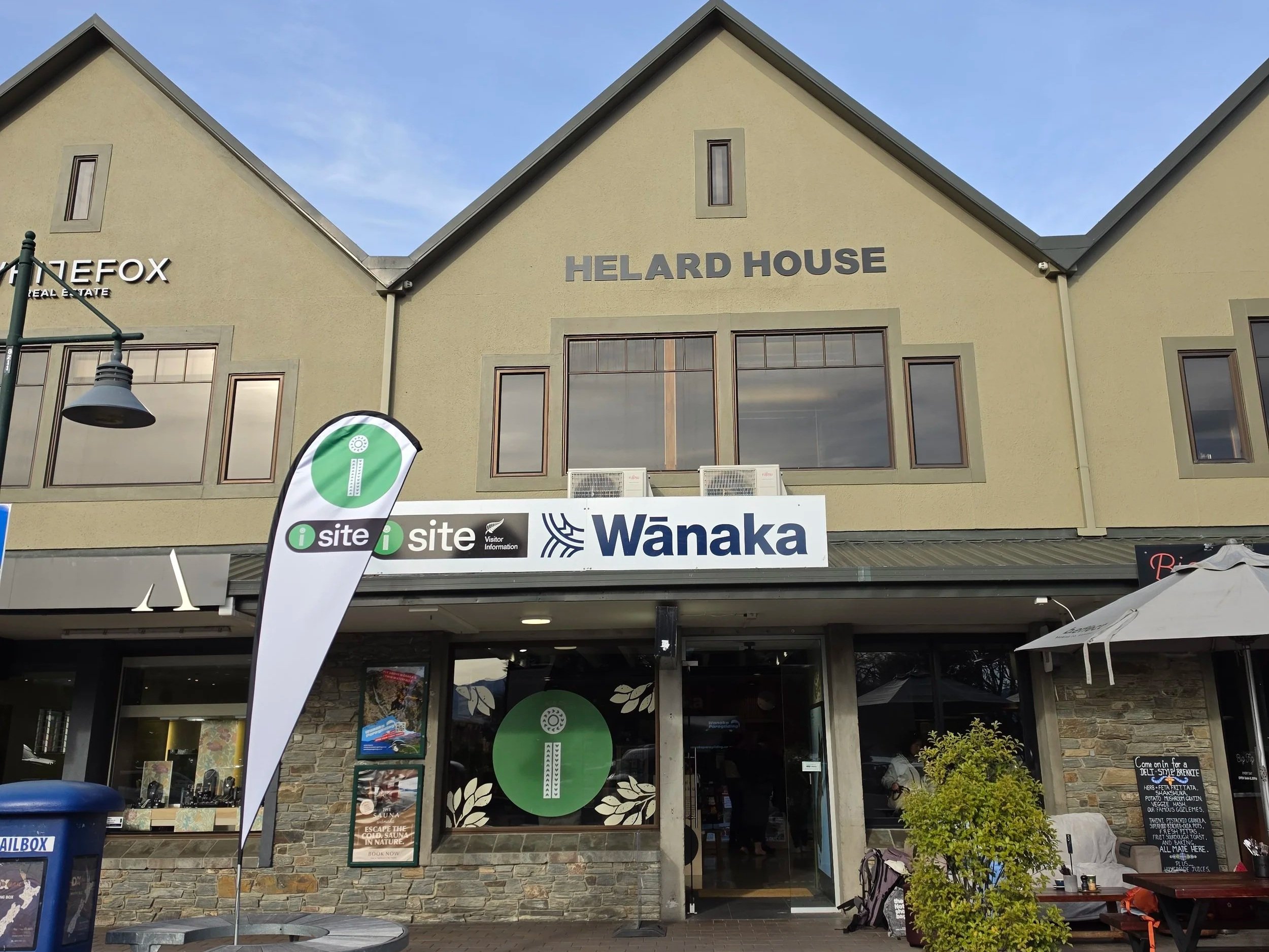 The building has signs for Helard House, Wanaka, and a yoga studio. There are flags, outdoor seating, a trash bin, and greenery in front of the establishment.