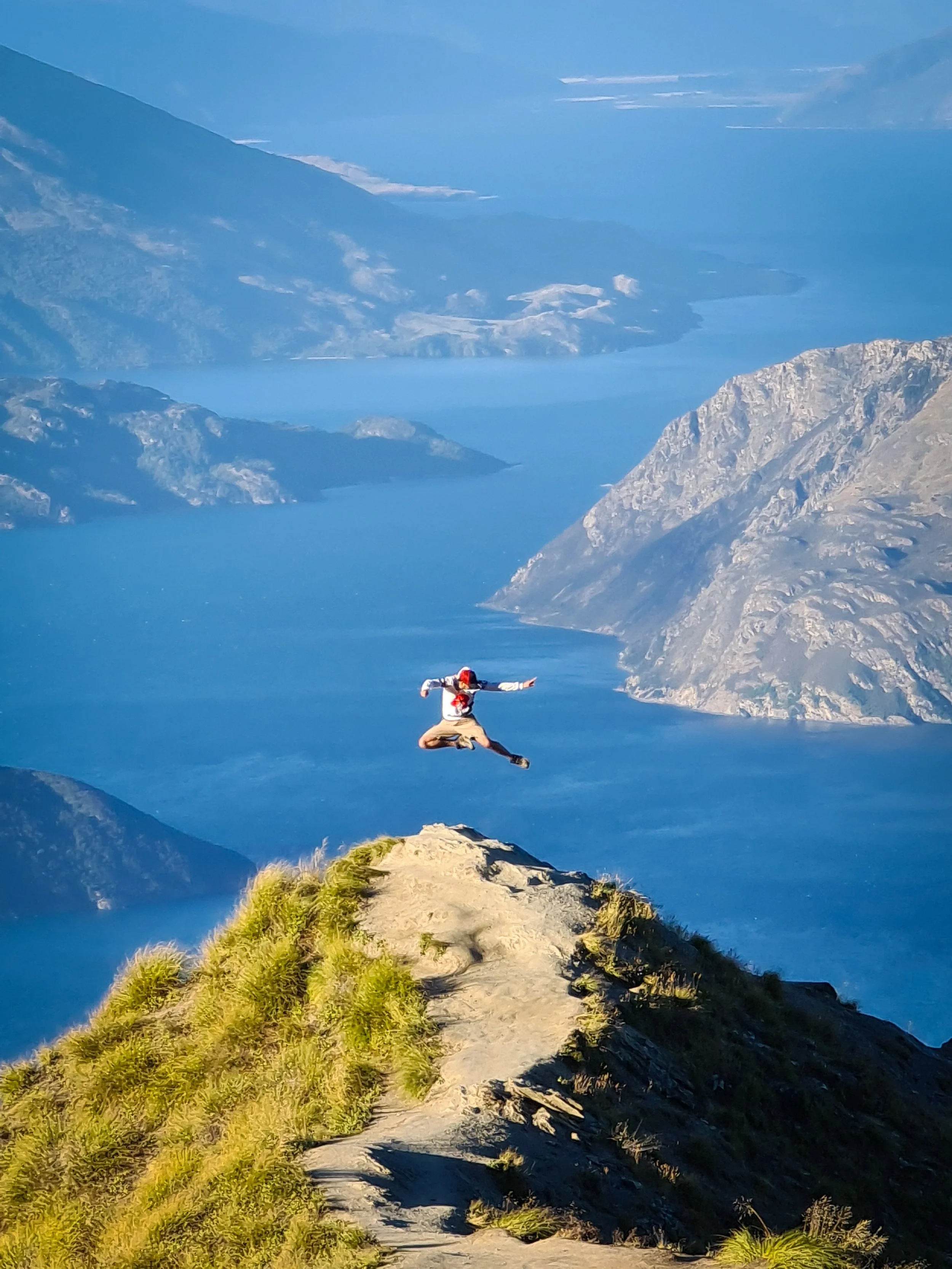 A person wearing a white shirt, beige pants, and a red hat is jumping with arms and legs outstretched on a mountain trail near a steep, grassy slope, with a deep blue lake and snow-capped mountains in the background.