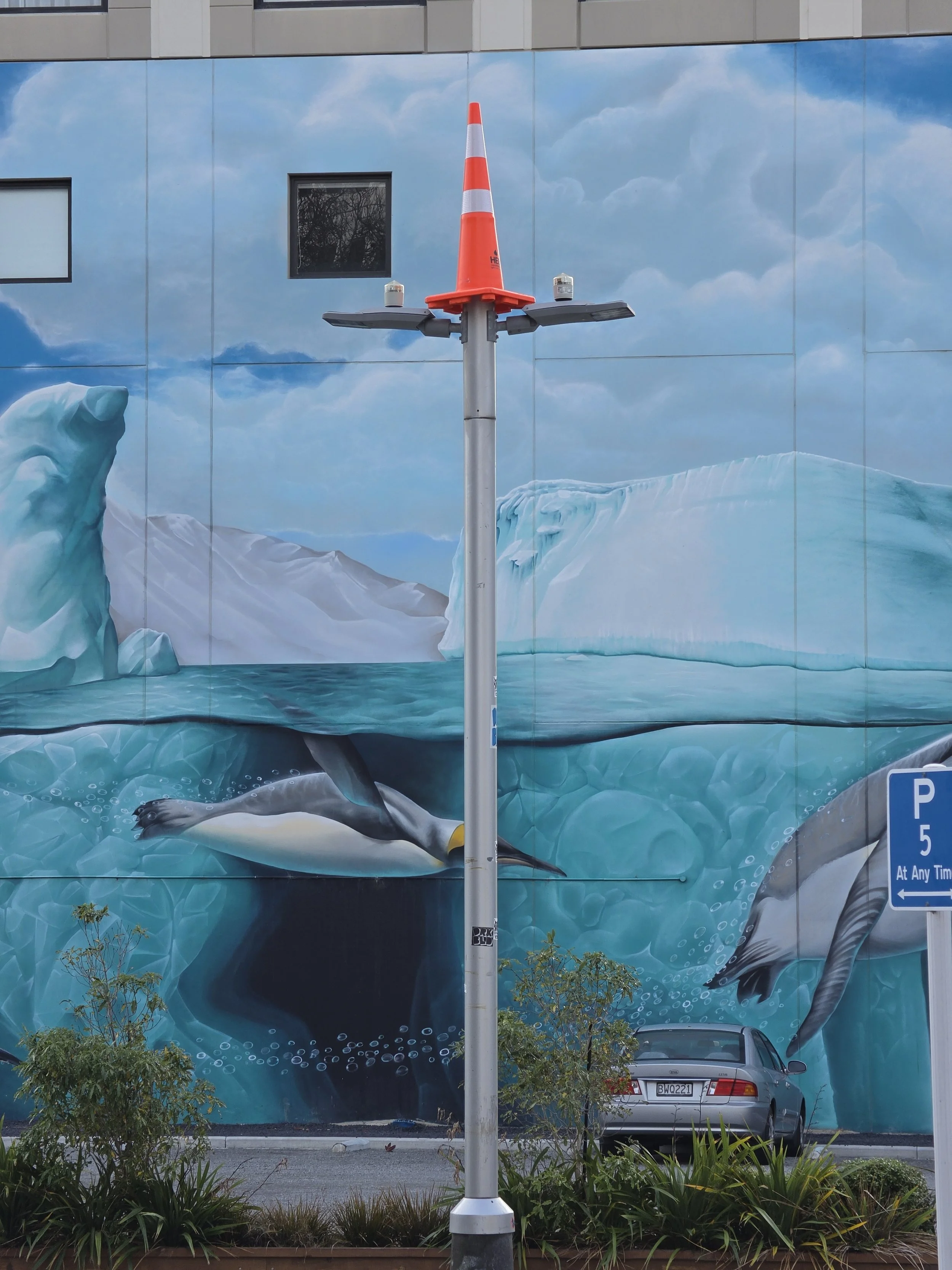 Large mural of an iceberg and a penguin on a building wall with a bright blue sky and clouds. A parking lot scene with a silver car and a parking sign is visible in front of the mural. A streetlight pole with a traffic cone on top is positioned in the center of the image.