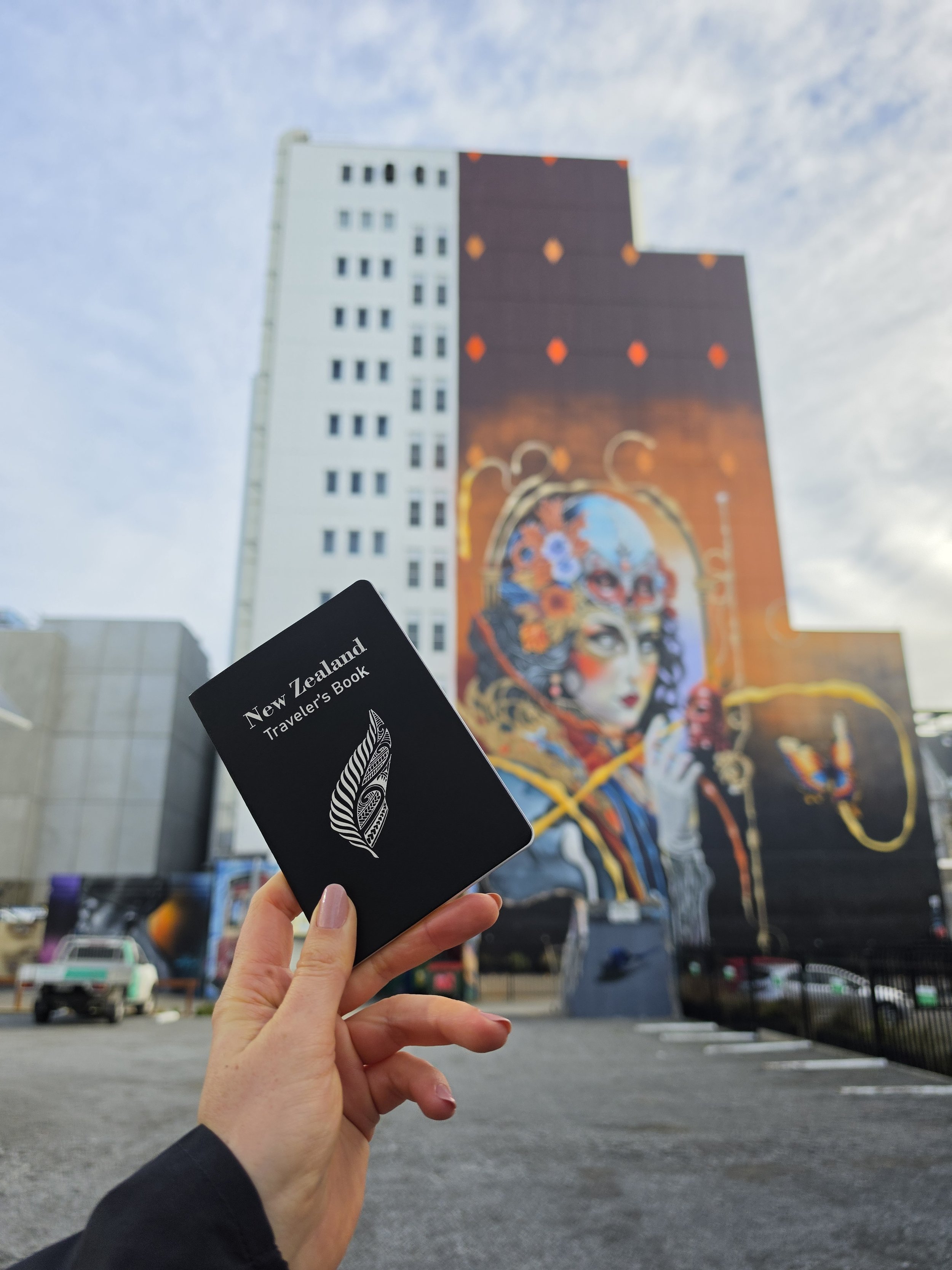 A person holding a black card that says 'New Zealand Traveler's Book' in front of a city street with cars and a tall building featuring colorful street art of a woman's face.