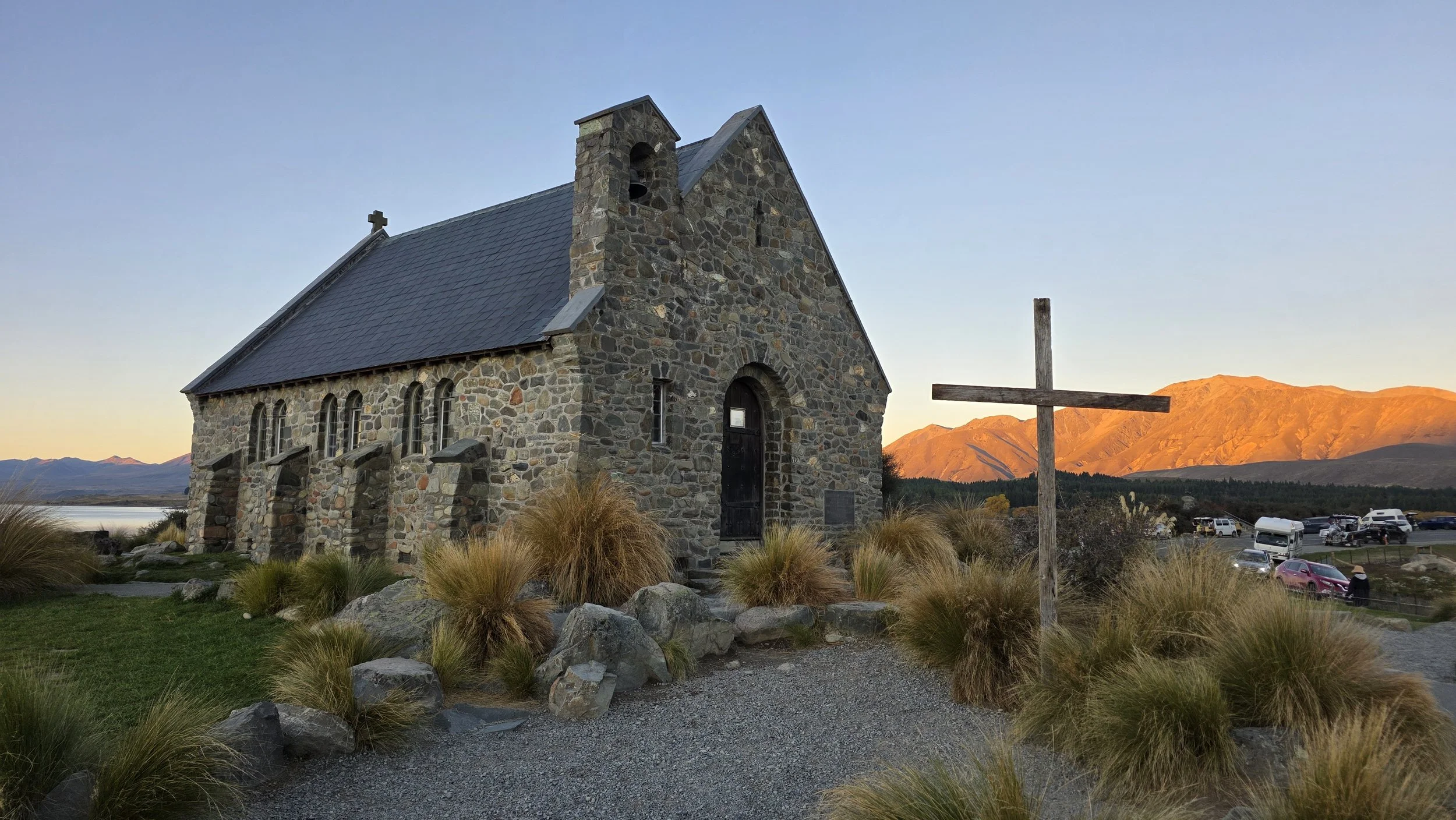 A small stone church with a steeple, surrounded by rocks and tall grasses, with mountains and a parking lot in the background during sunset.