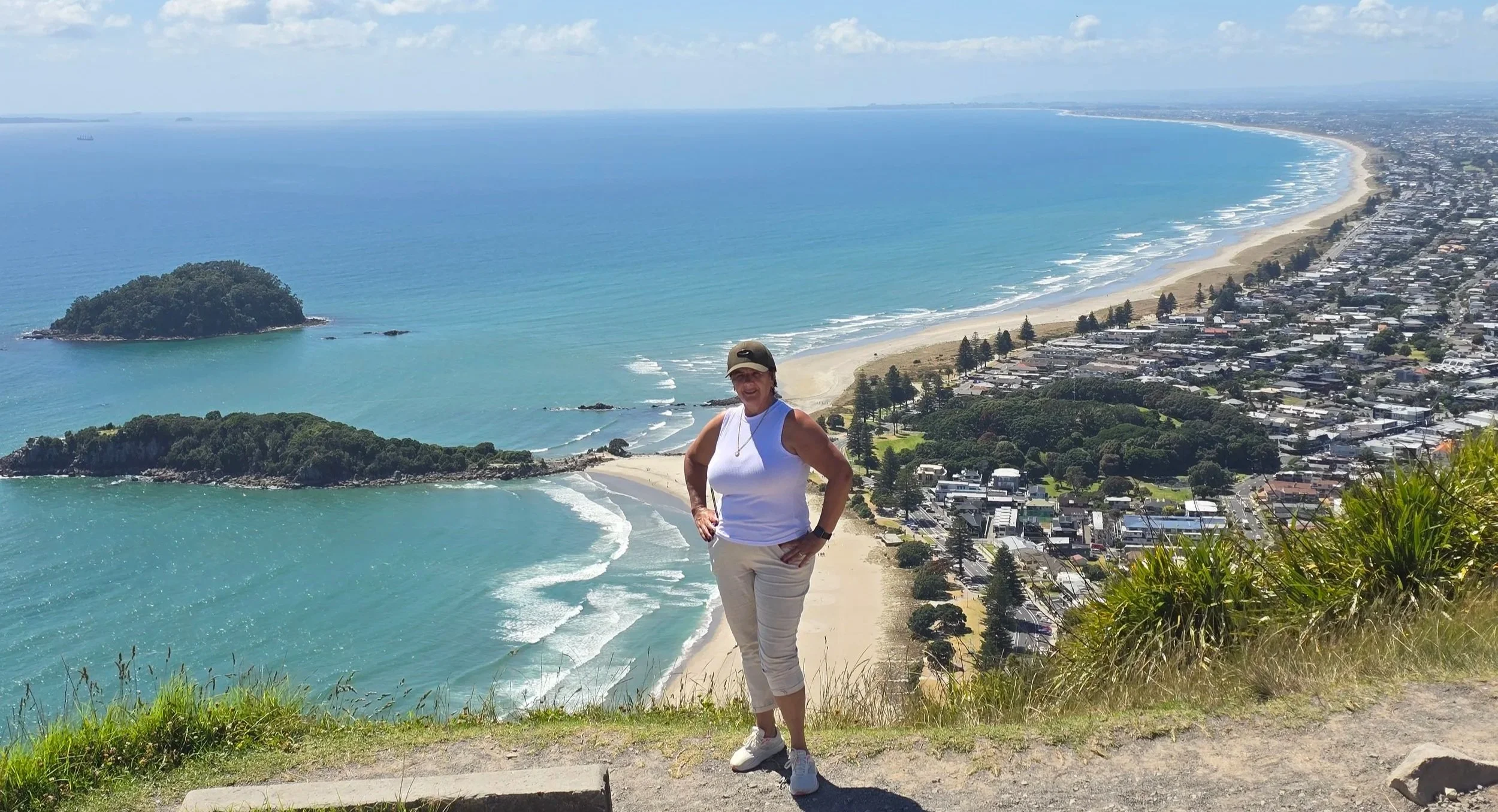An older woman wearing a white sleeveless top, light-colored pants, and a beige cap standing on a hillside overlooking a coastal town with a beach, ocean, and greenery.