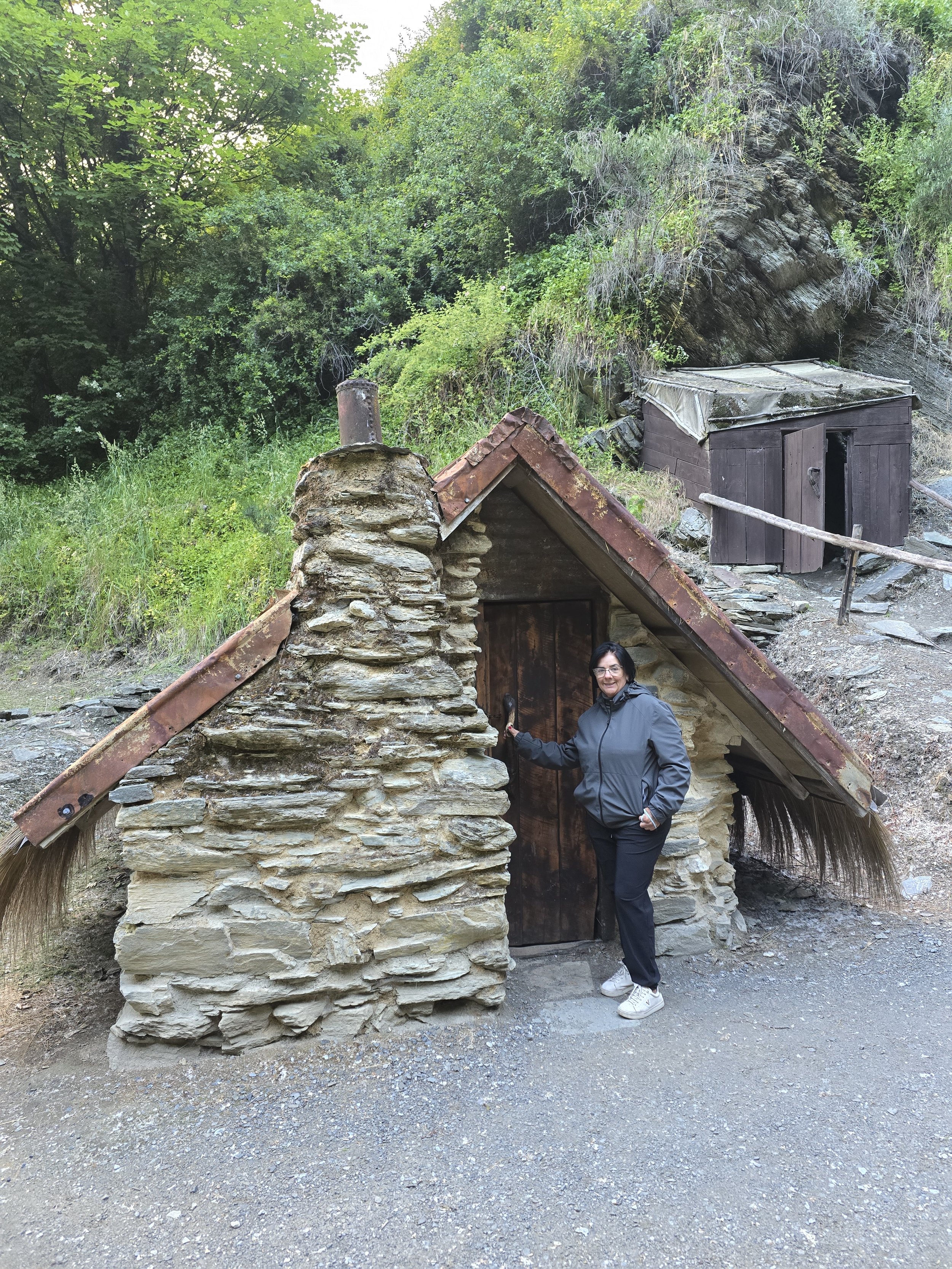 A woman wearing a gray jacket and black pants standing at the entrance of a small, rustic stone and wood cabin in a wooded area with green trees and rocks in the background.