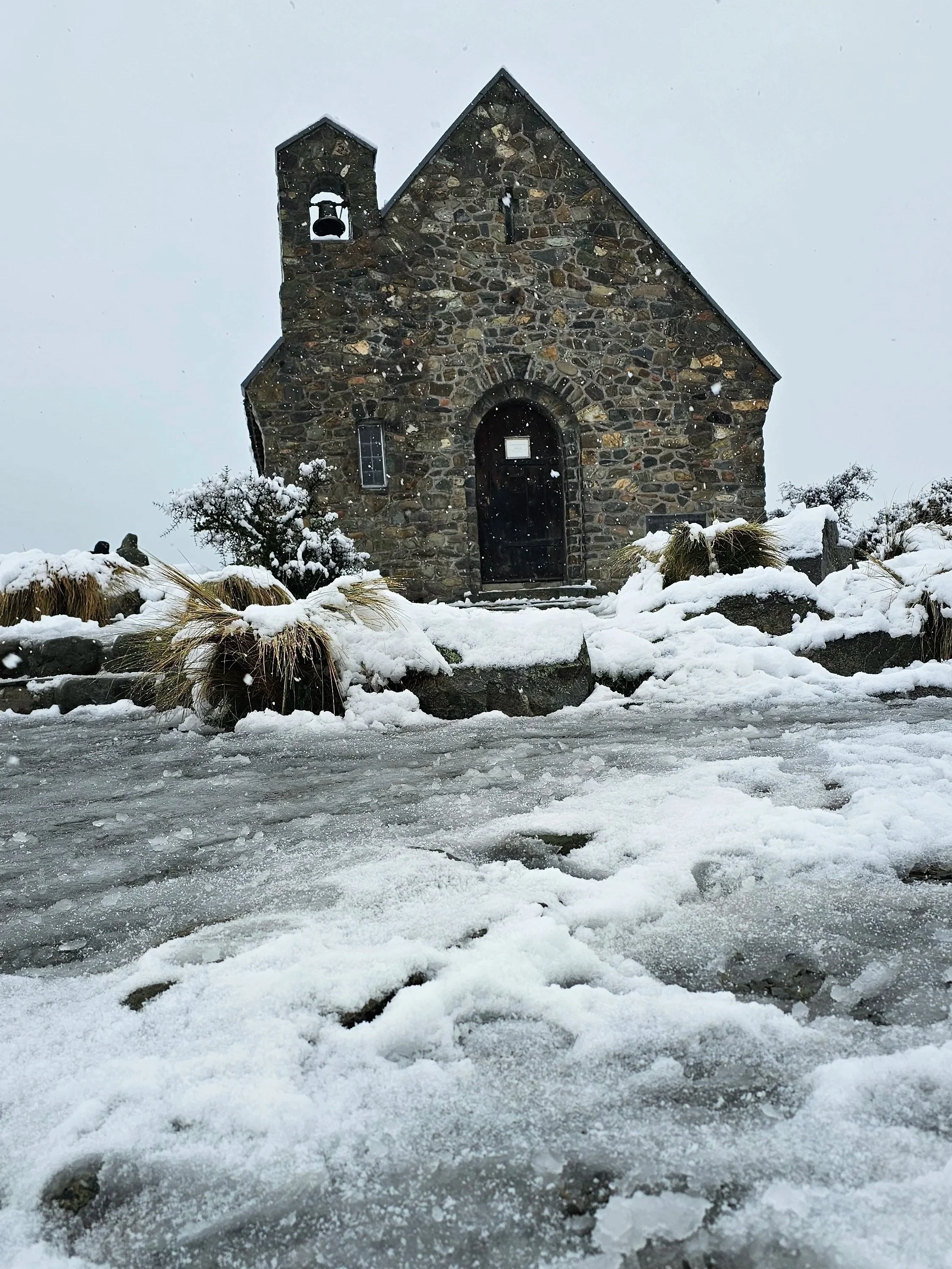 Stone church surrounded by snow with footprints on icy ground and snow-covered bushes in the foreground.