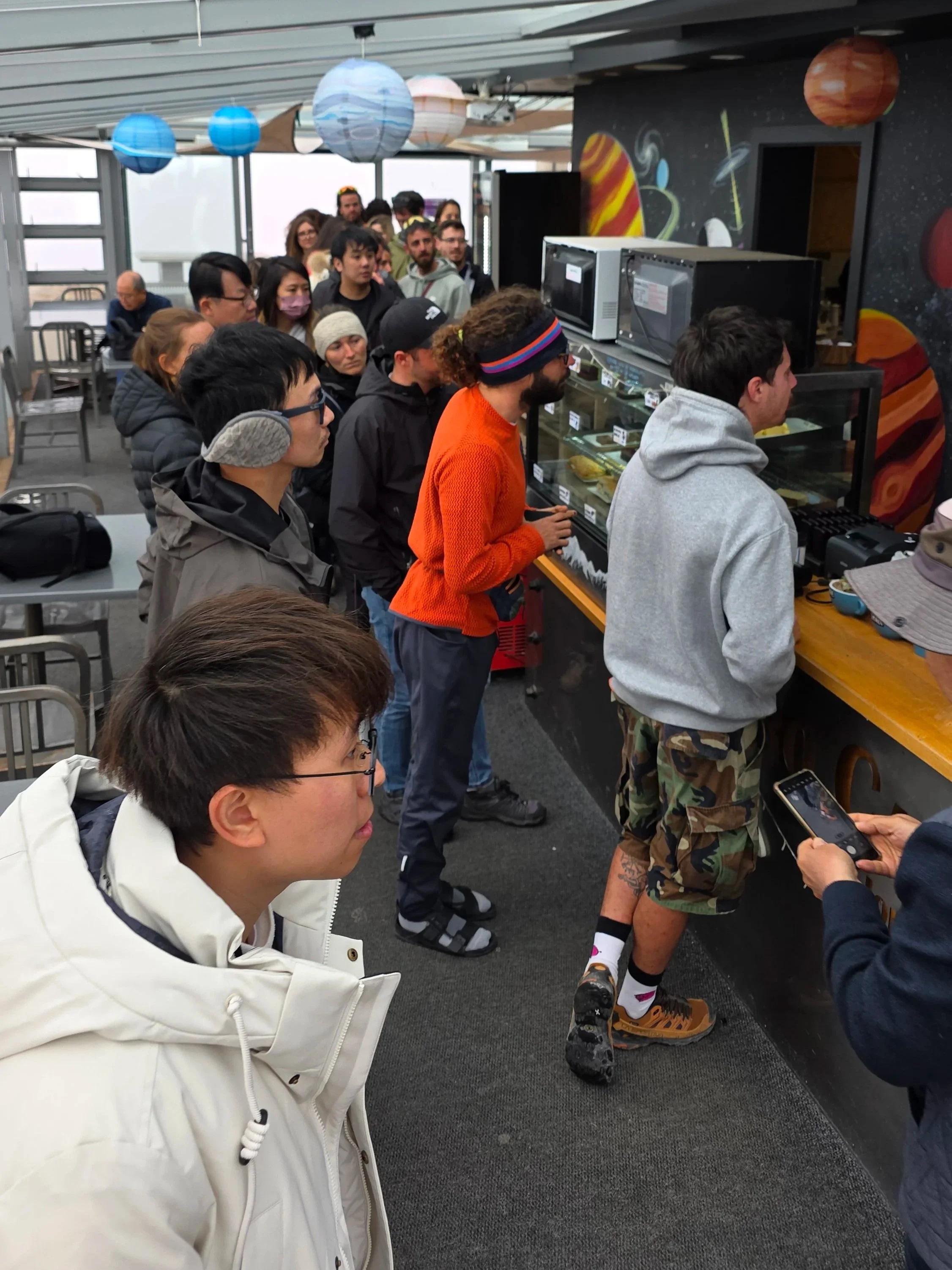 People standing in line, waiting to buy food at a cafeteria counter with space-themed decorations on the wall and hanging lanterns resembling planets and stars.