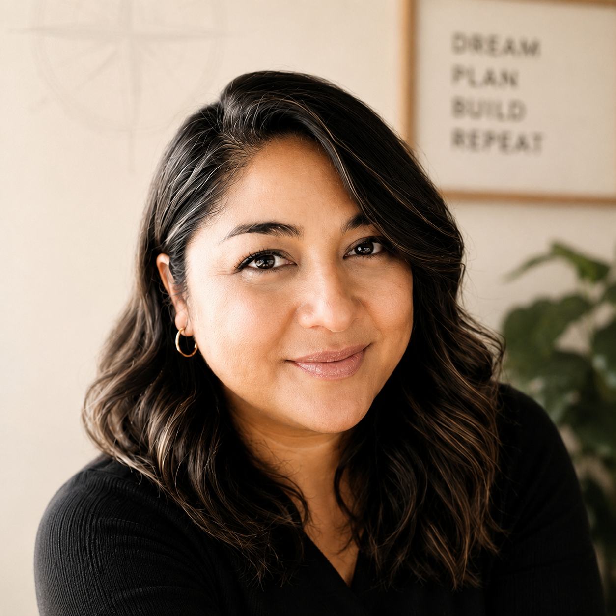 Close-up portrait of a woman with shoulder-length wavy dark hair with highlights, wearing hoop earrings and a black top, smiling softly at the camera. In the background, there is a blurred motivational poster with phrases such as "DREAM," "PLAN," "BUILD," and "REPEAT," along with some greenery.