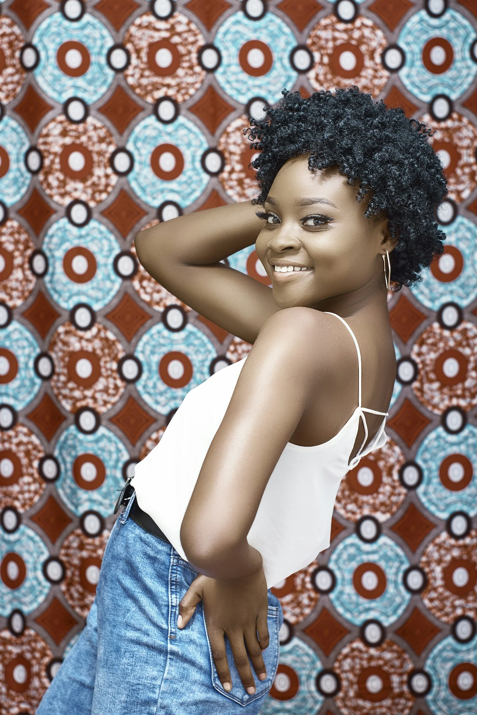 A young woman with short curly black hair smiling and posing with her hand behind her head, wearing a white sleeveless top and blue jeans, standing against a colorful geometric patterned background.