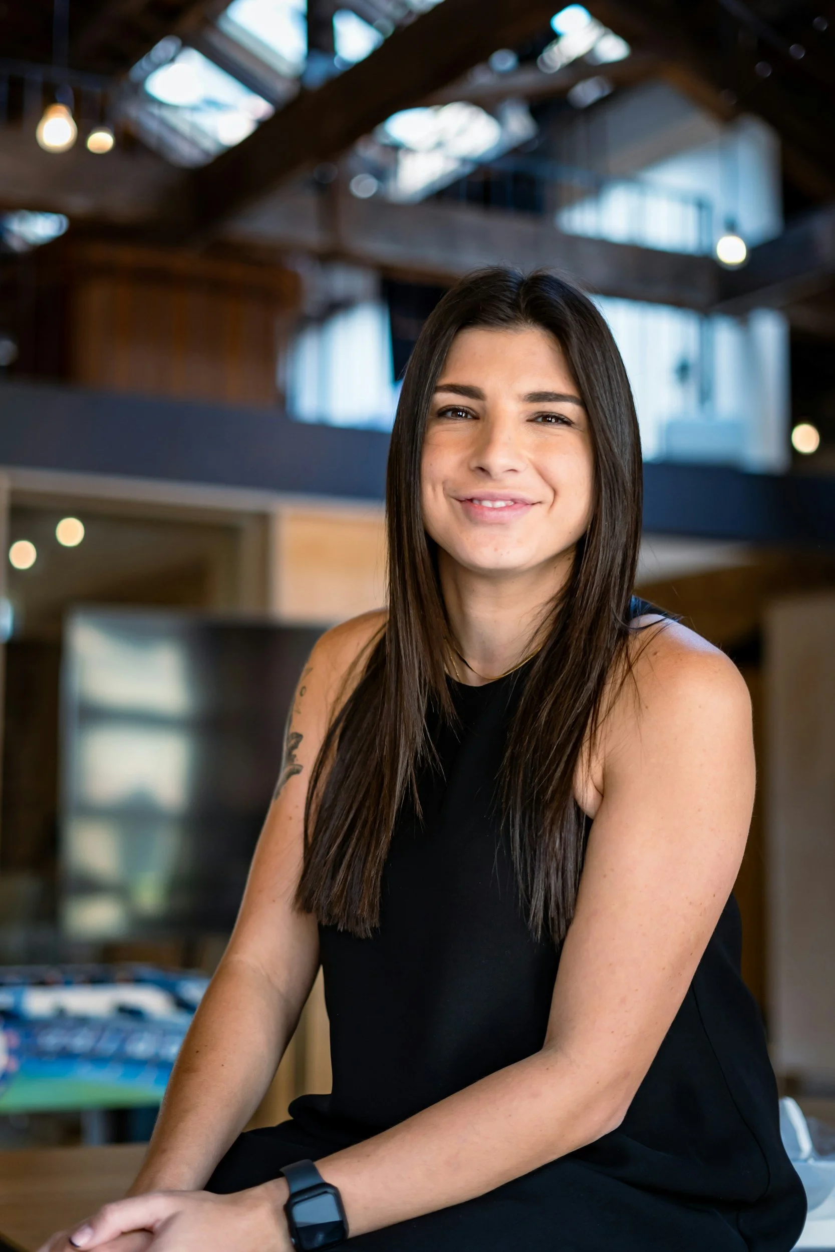 A young woman with long dark hair, wearing a sleeveless black top, sitting indoors in a modern, rustic space with wooden beams and large windows, smiling at the camera.