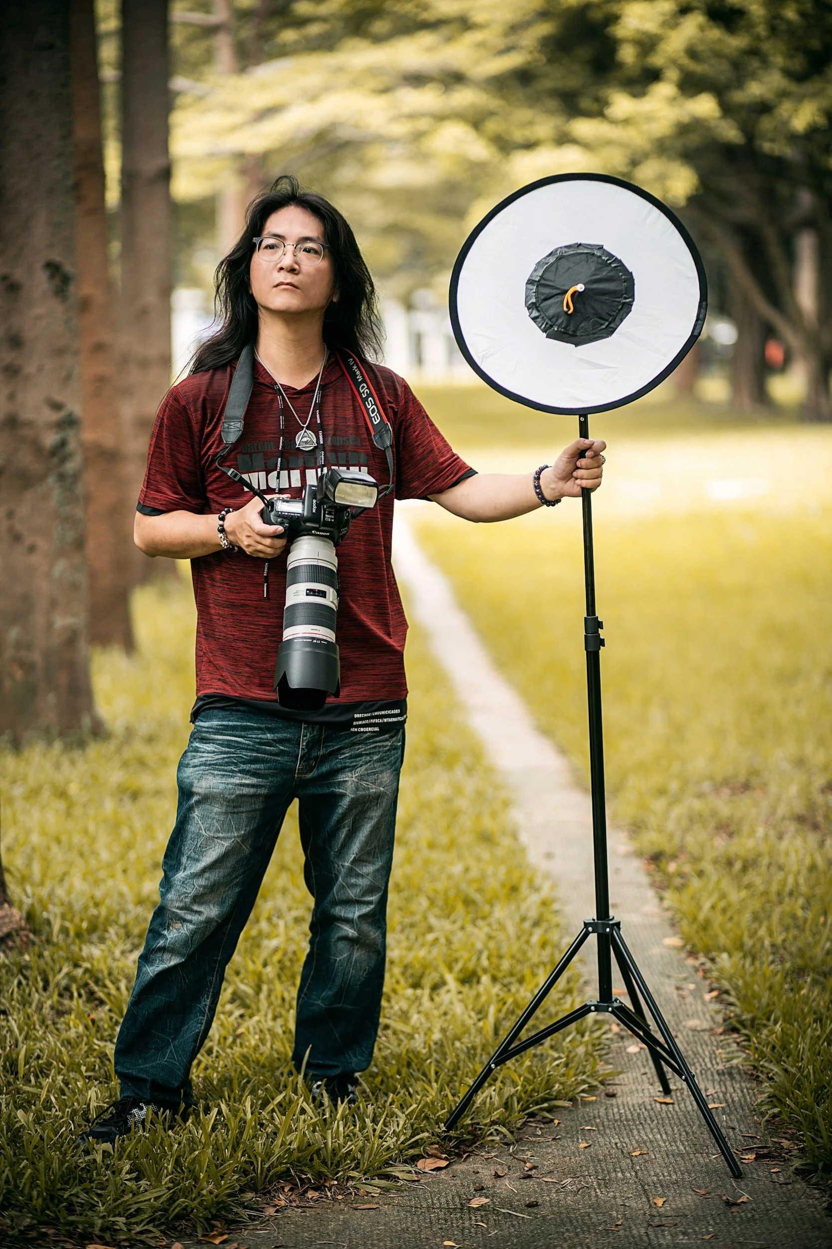 A person with long dark hair, glasses, and a camera around their neck, standing outdoors on a grassy path surrounded by trees, holding a large circular reflector on a stand.