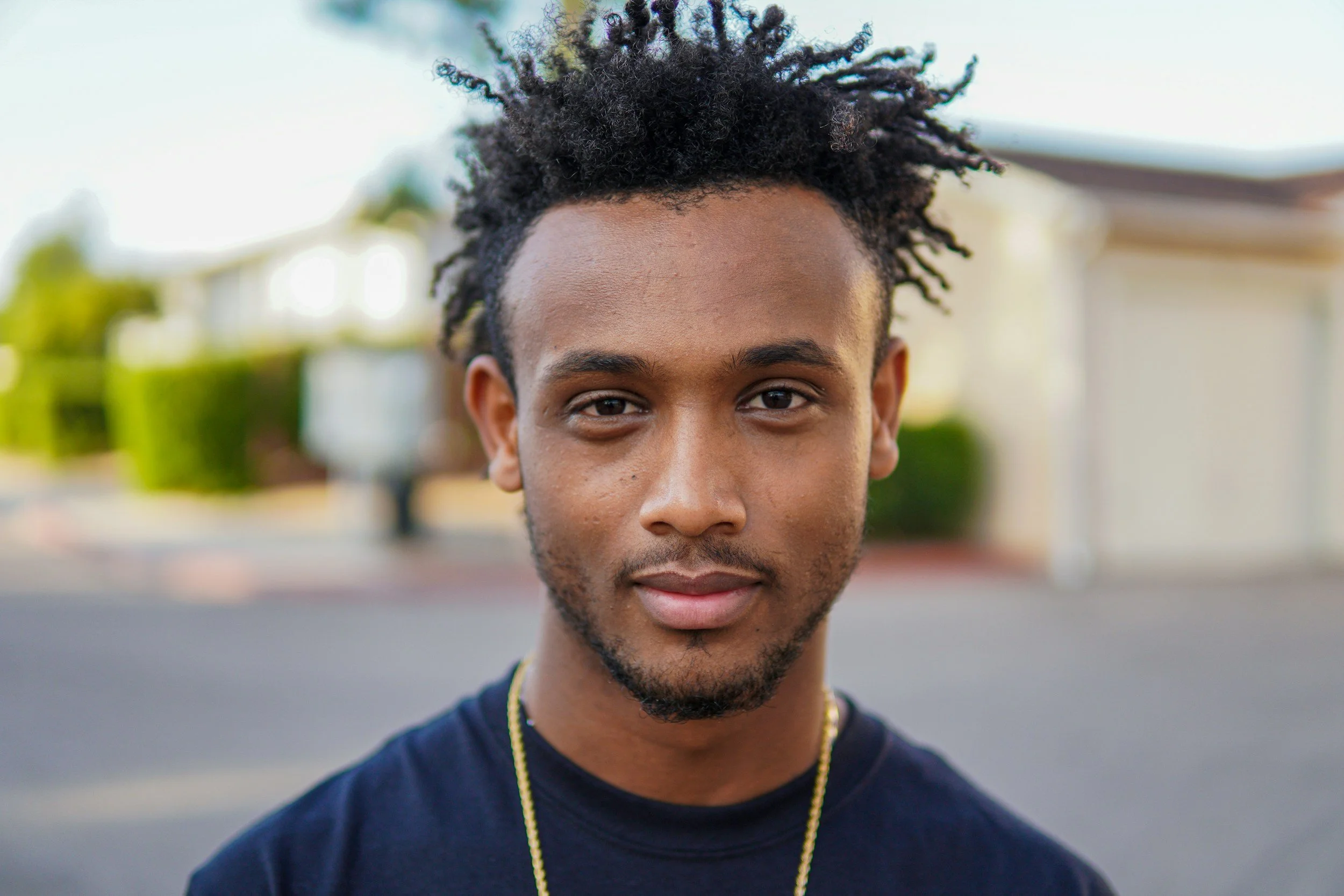 Close-up portrait of a young man with dark, curly hair and a slight beard, standing outdoors in a suburban neighborhood with houses and greenery in the background.