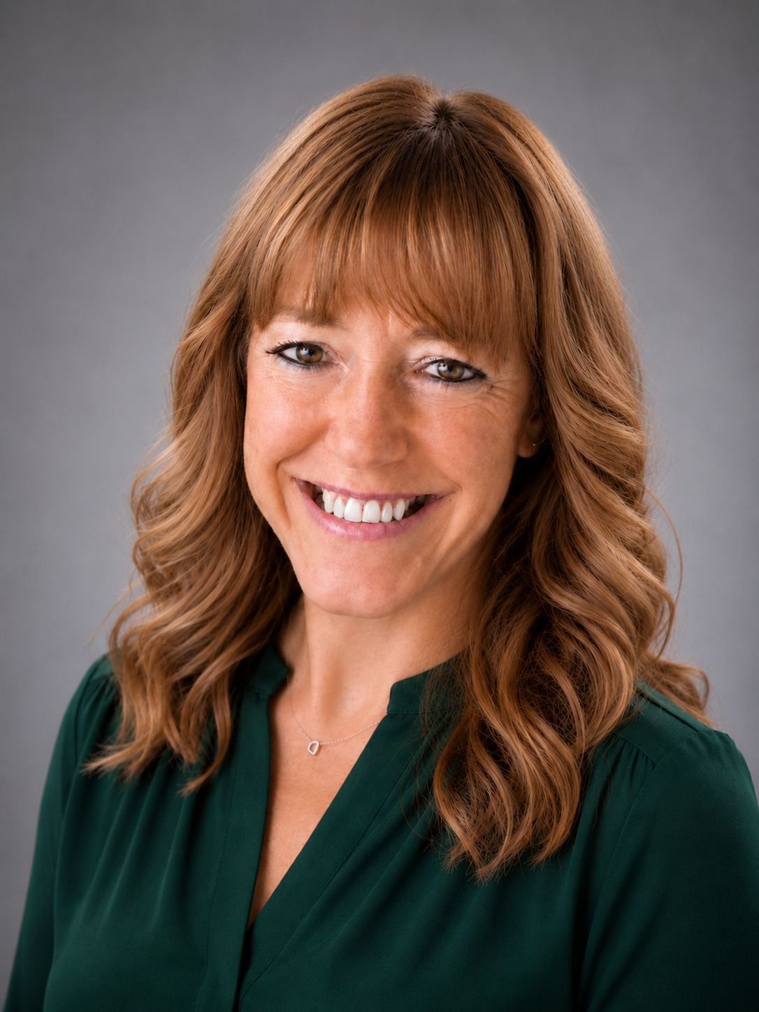 A woman with wavy red hair, smiling, wearing a dark green blouse and a silver necklace with a heart-shaped pendant, against a gray background.