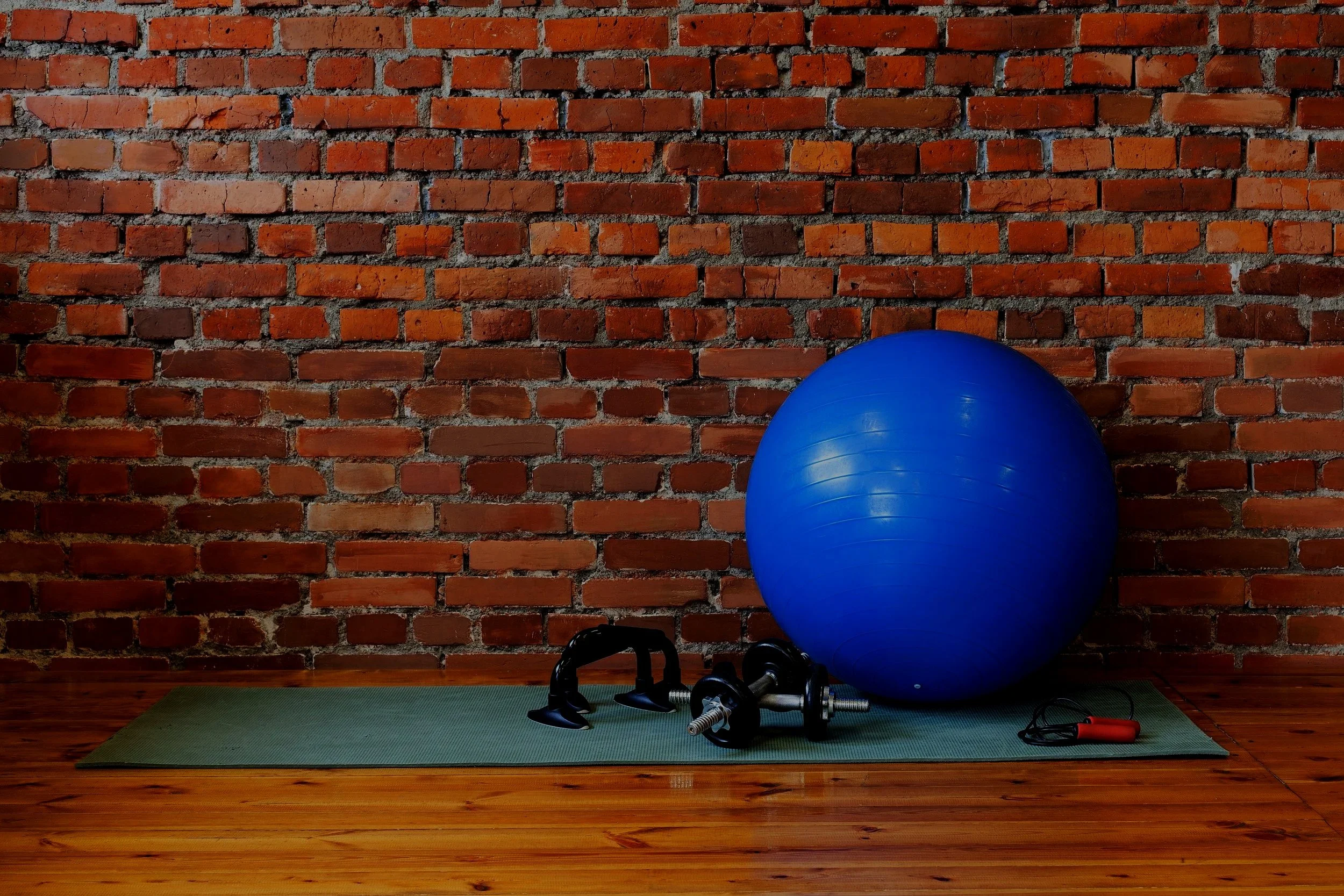 Fitness equipment including a large blue exercise ball, kettlebell, dumbbell, jump rope, and resistance band on a green exercise mat, set against a brick wall with wooden flooring
