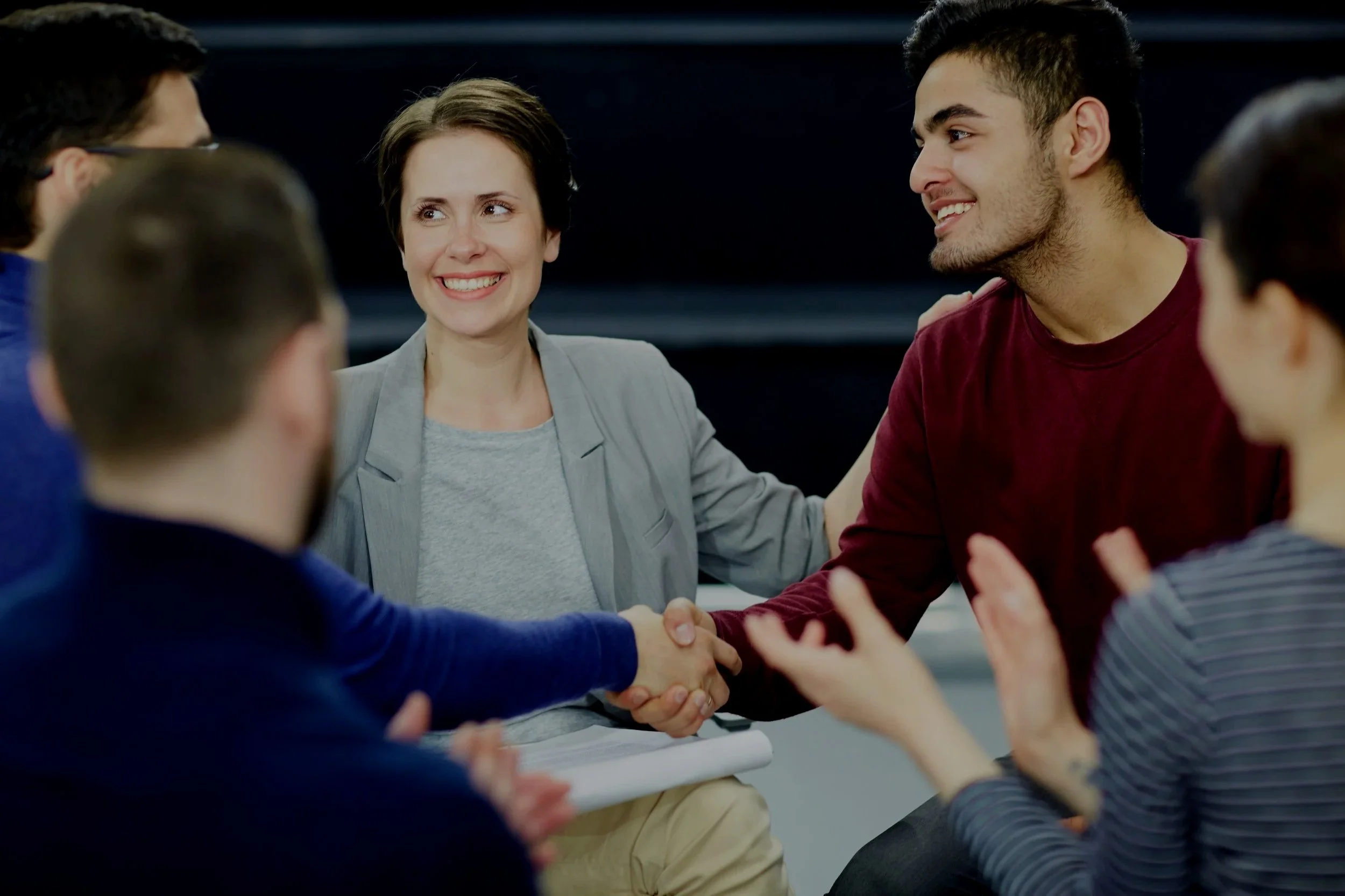 A diverse group of people in informal attire sitting together in a circle, engaging in a discussion, with one woman smiling and a young man shaking hands during the conversation.
