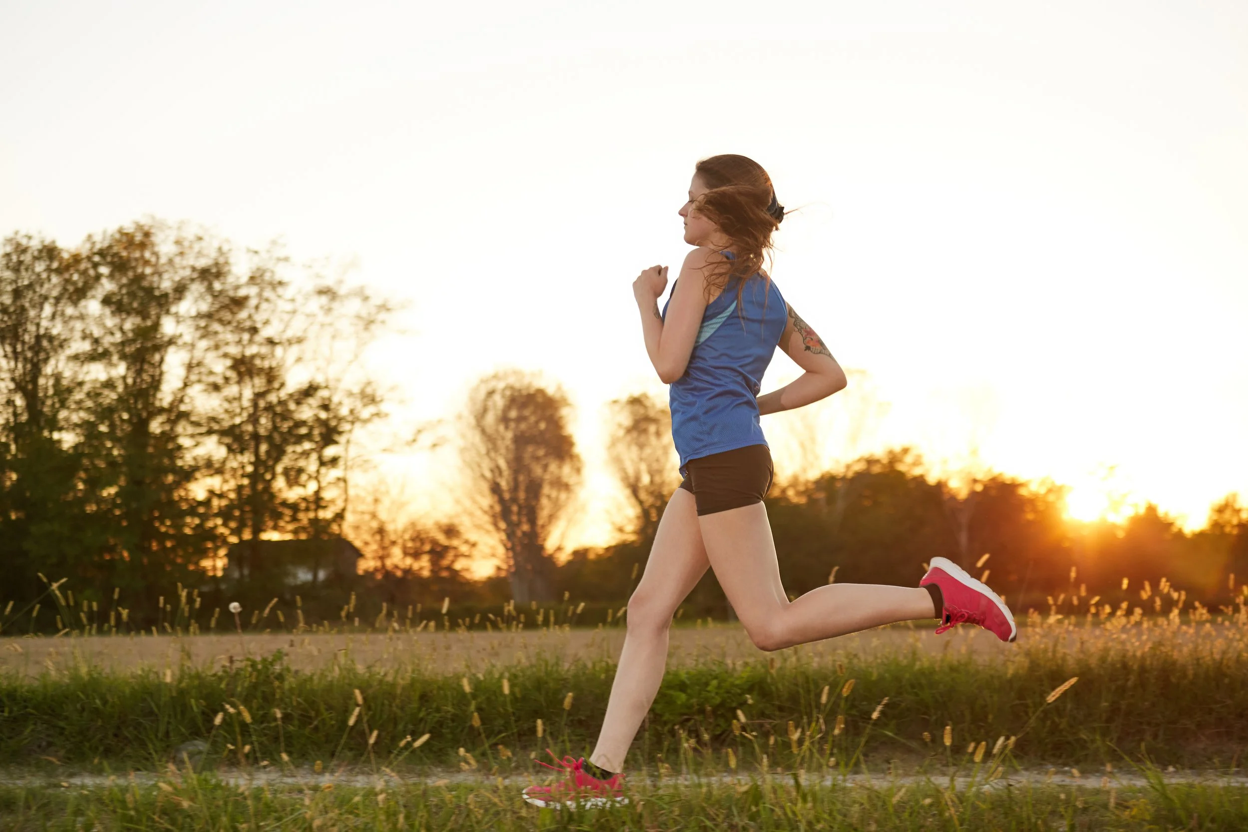 A woman running outdoors at sunset, wearing a blue athletic tank top, black shorts, and red running shoes, with trees in the background.