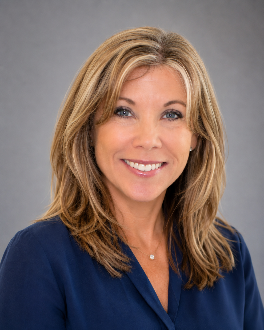 Headshot of a smiling woman with shoulder-length blonde hair, wearing a navy blue top and a small necklace, against a gray background.
