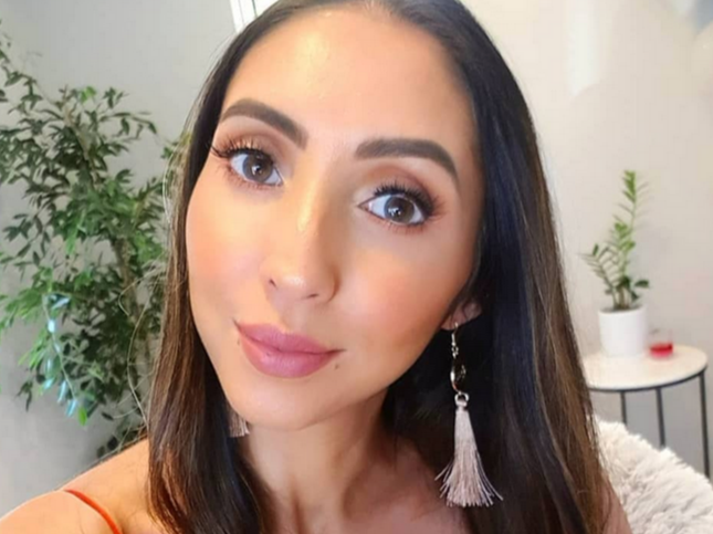 Close-up of a young woman with dark hair, wearing earrings and makeup, smiling in front of indoor plants and a small table.