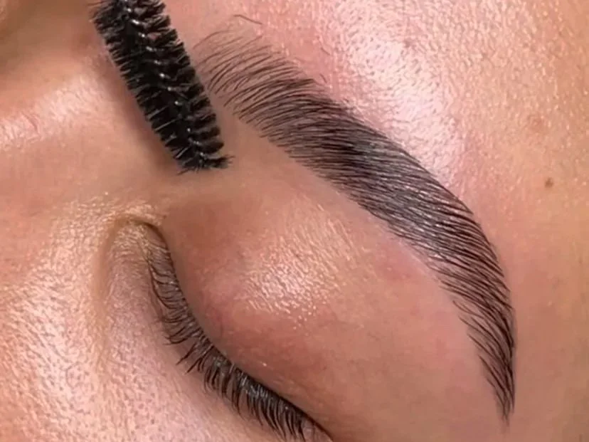 Close-up of a person's eyebrow being brushed with a spoolie brush, with the person's eye closed.