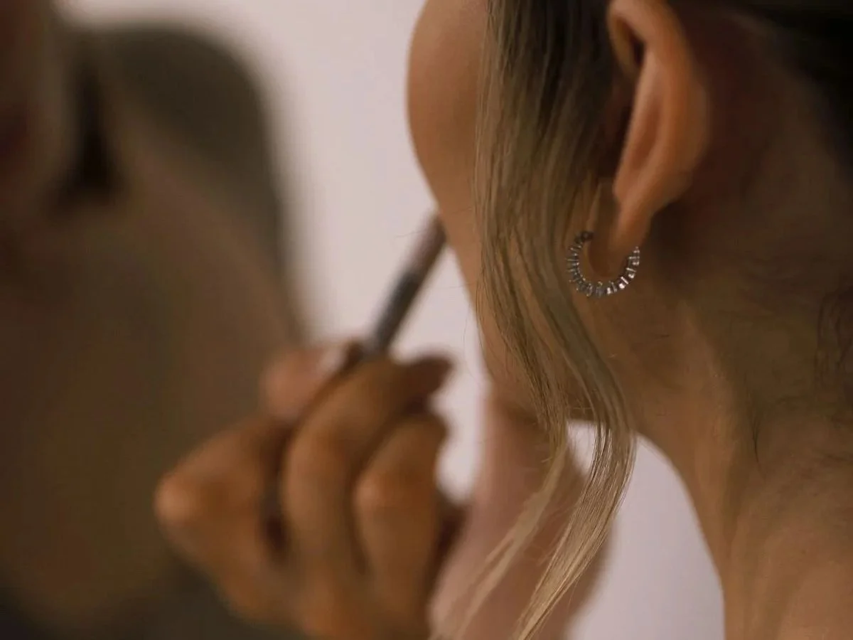 Close-up of a person applying makeup with a brush, wearing an earring with a circular design, and with wavy hair.