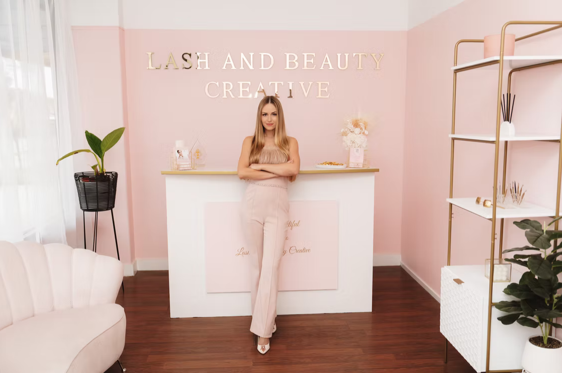 A woman standing with arms crossed in a pink and white beauty salon reception area with plants and shelves.
